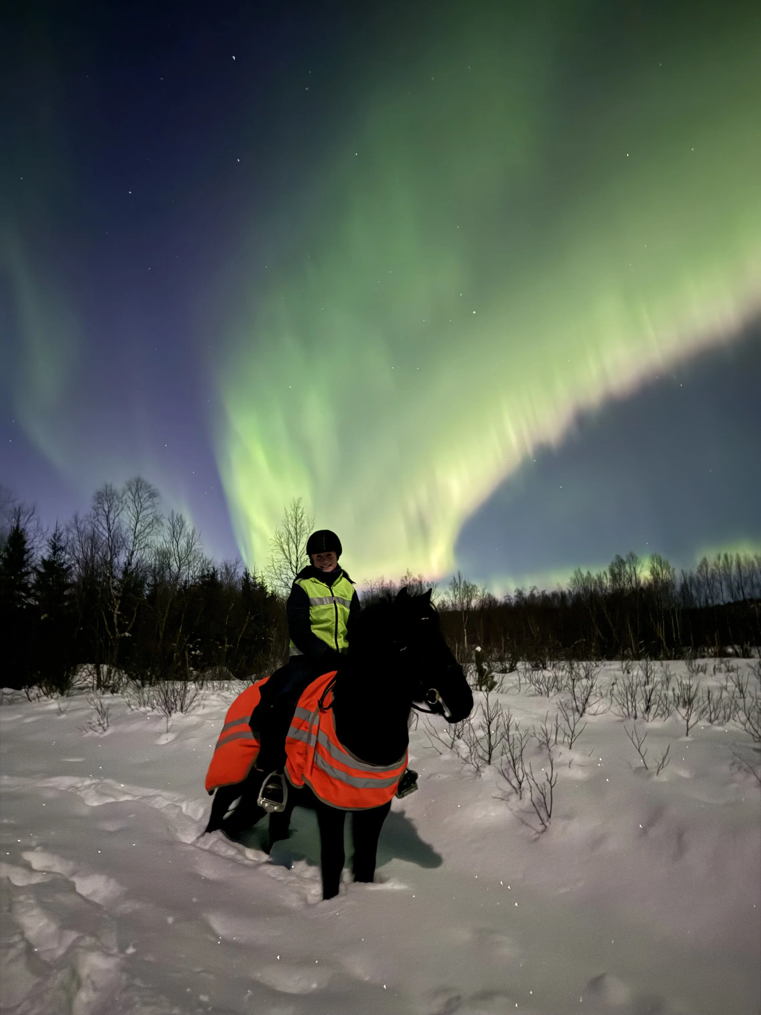 Person som rir på hest i snøen under nordlyset, iført vinterklær og sikkerhetsutstyr som refleksvest og hjelm.