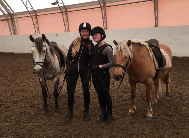 To unge kvinner i rideklær står med hester i en innendørs ridehall hos Stall Holand på Tverlandet i Bodø / Nordland, begge smilende og iført hjelm.