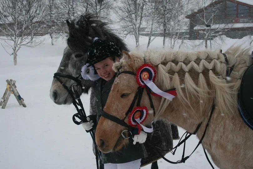 To hester og en jente utendørs i snøvær, begge med rosetter i halsene, og jenta smiler.