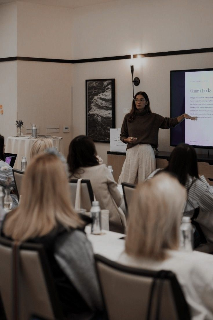 A woman in a brown sweater and cream-colored pants presenting a slideshow to a group of women seated at tables in a conference room.