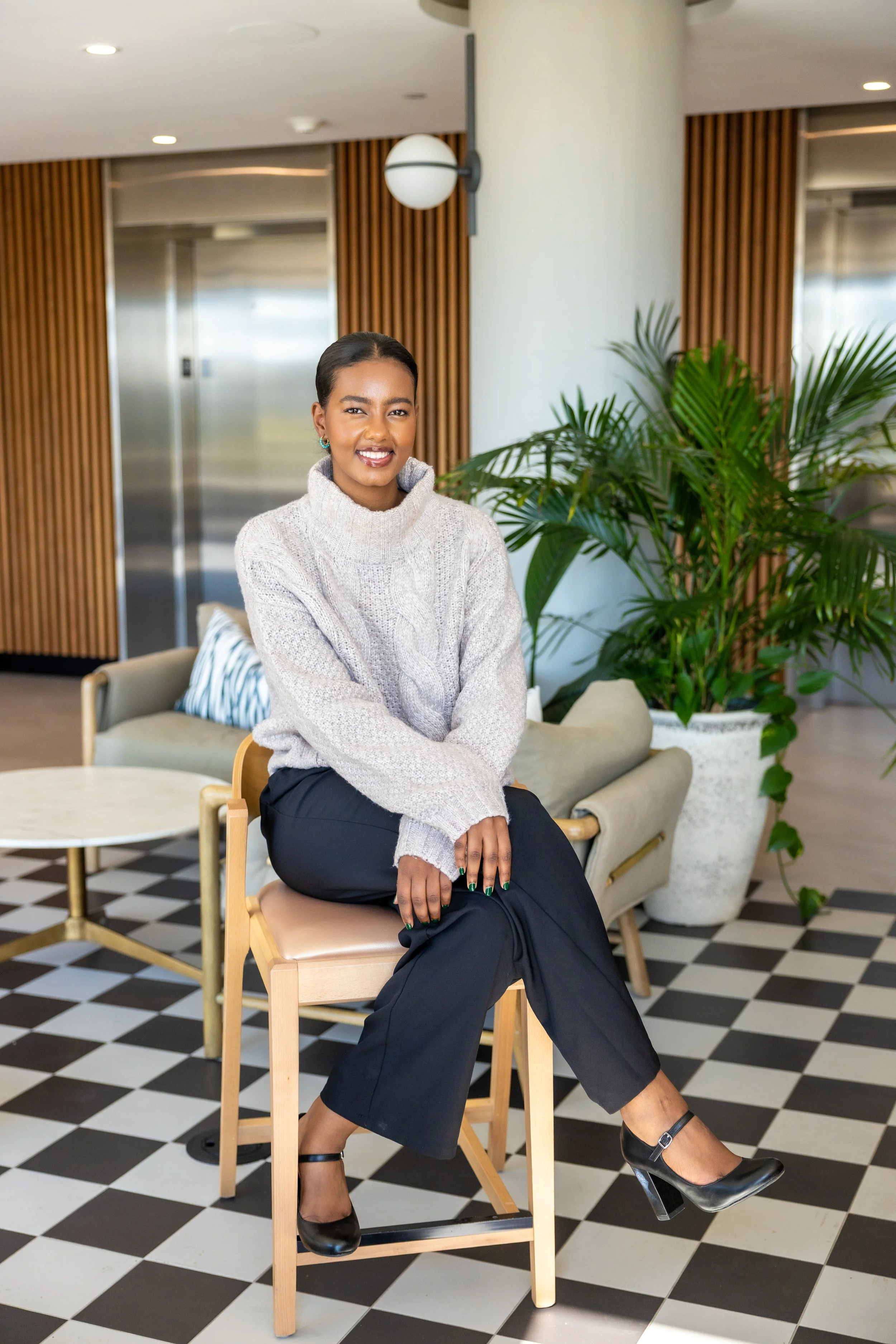 A smiling woman with dark hair tied back, wearing a gray knit turtleneck sweater, black pants, and black heels, seated on a wooden chair in a modern hotel lobby with black and white checkered floor, green indoor plants, and wooden wall paneling.