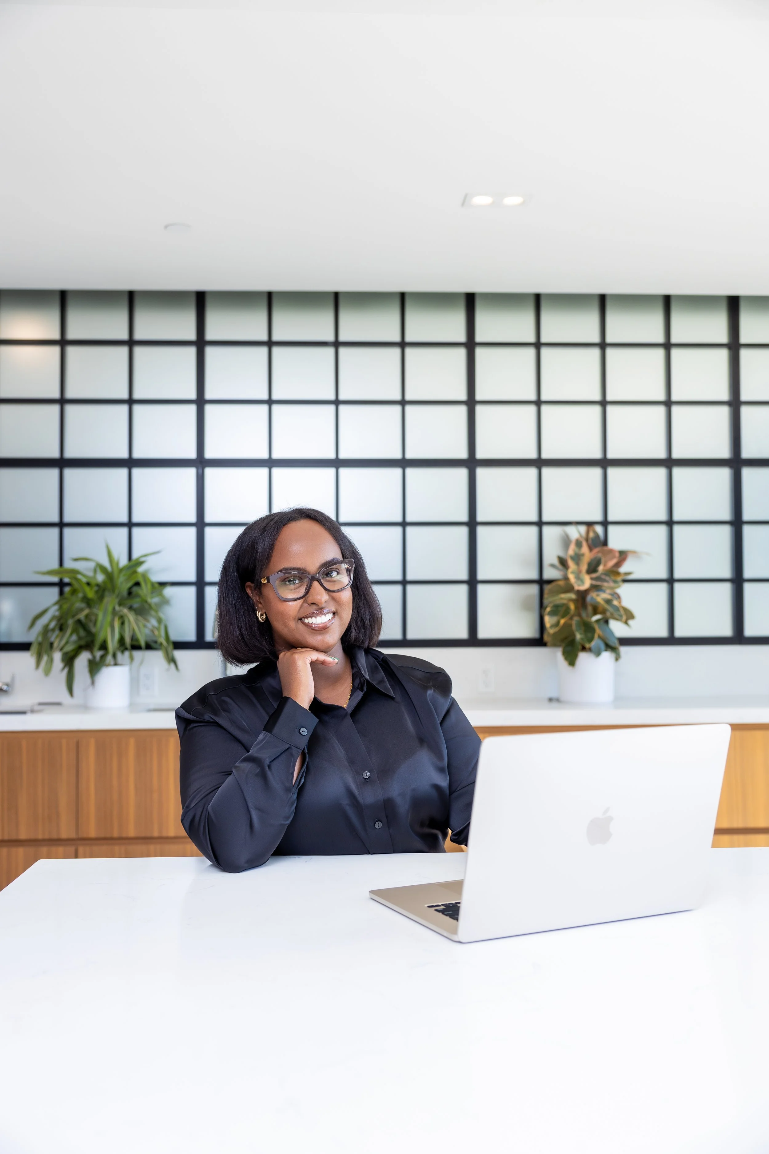 A woman with glasses sitting at a white desk with a laptop, smiling, in a modern office with plants and black grid wall decor.