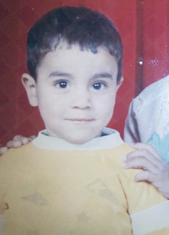 A child with short dark hair, wearing a yellow sweater, against a red background.