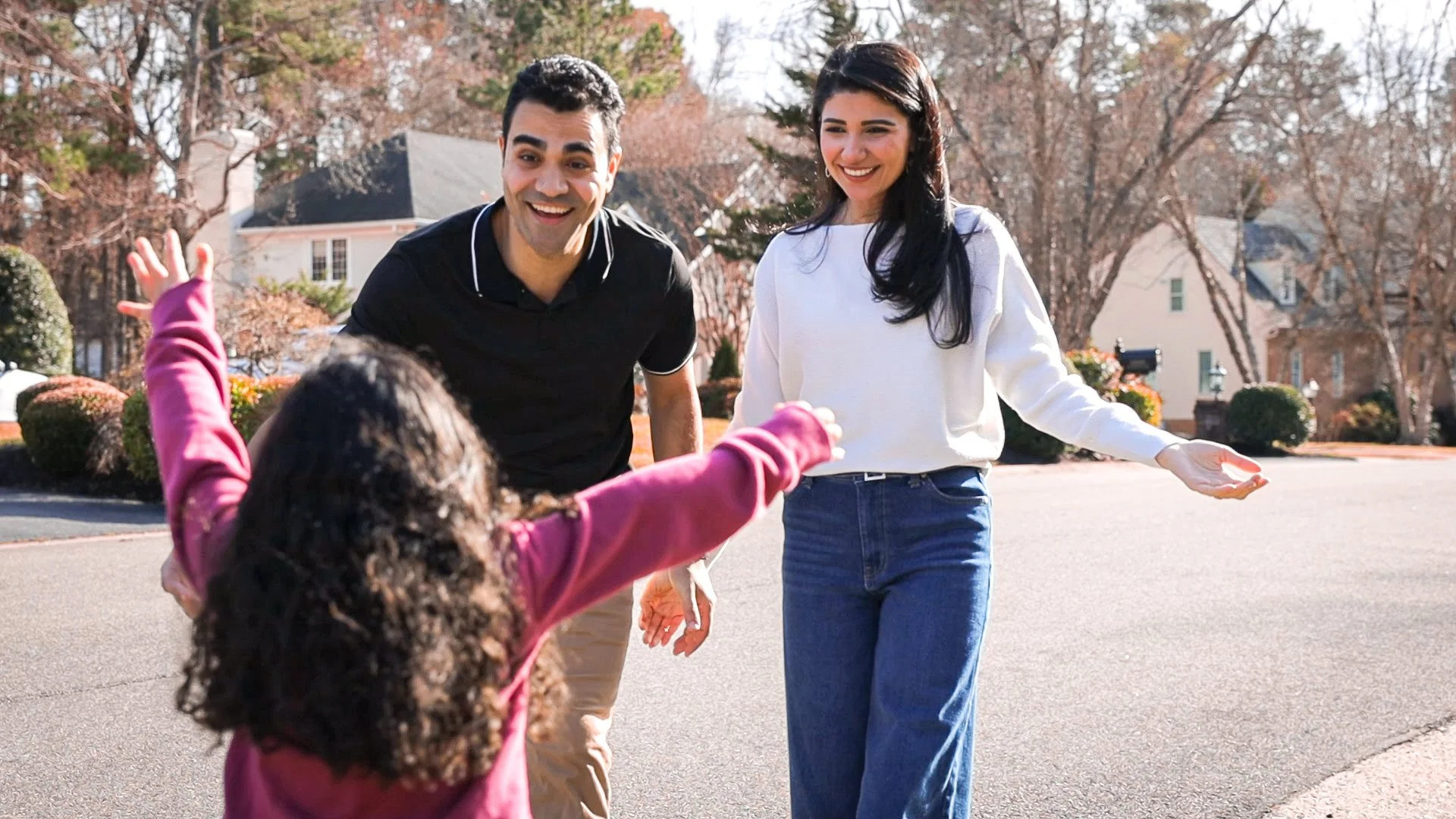 A happy couple welcoming a child running towards them in a suburban neighborhood.