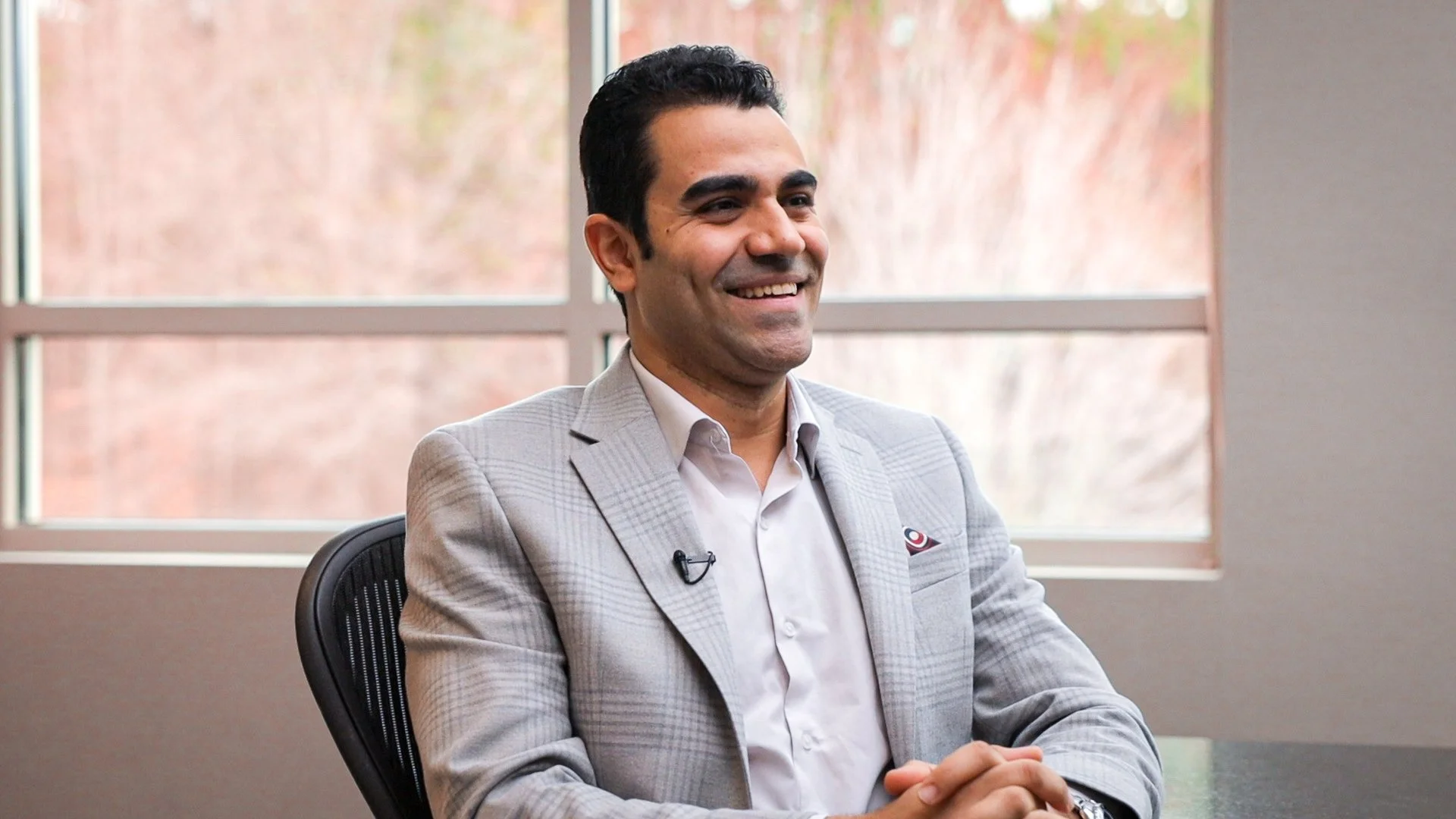 Milad Mikhail smiling in gray suit sitting at a table indoors with windows in the background.
