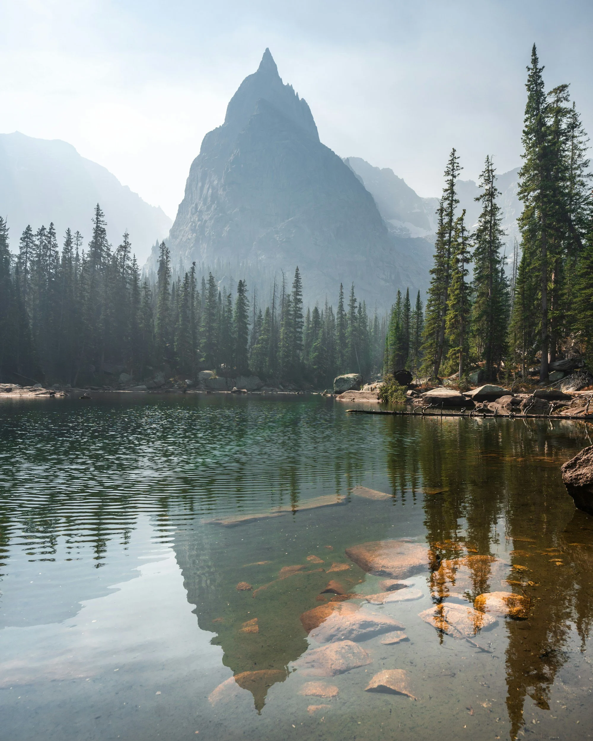 Tall mountain with sharp peak, surrounded by pine trees, reflecting in a calm lake with rocks along the shore.