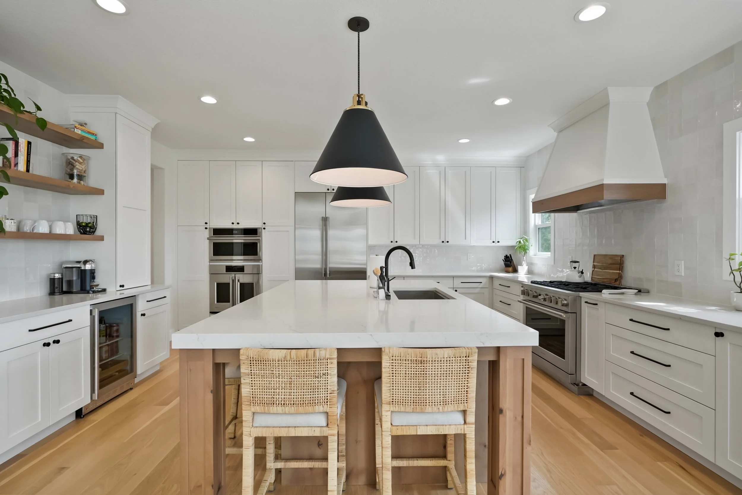 Modern white kitchen with black pendant lights over a central island, open shelving with books and bowls, stainless steel appliances, wood flooring, and natural light.
