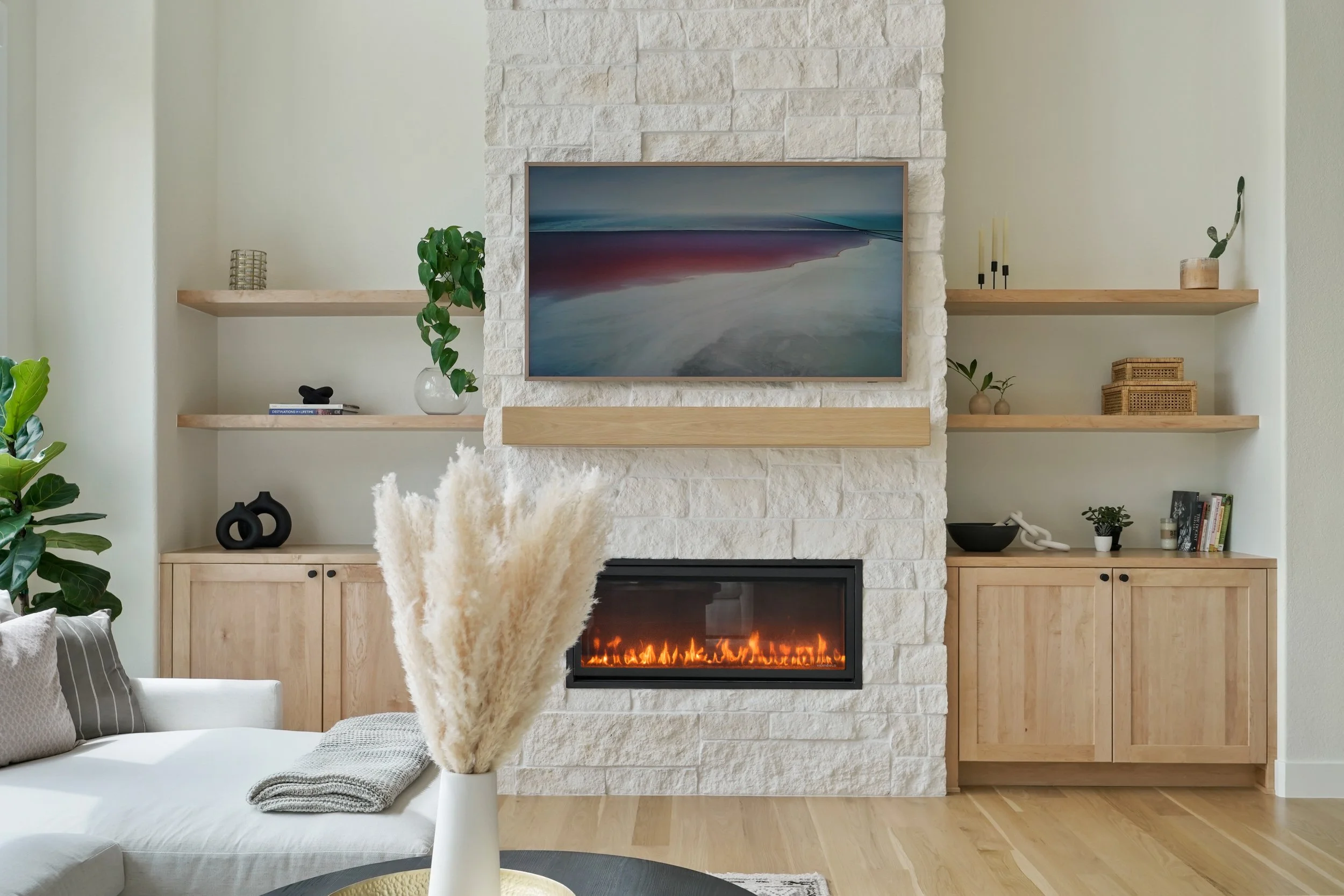 Living room with a fireplace, wall-mounted TV, wooden shelves with decorative items, plants, and a white sofa with throw pillows and a blanket.