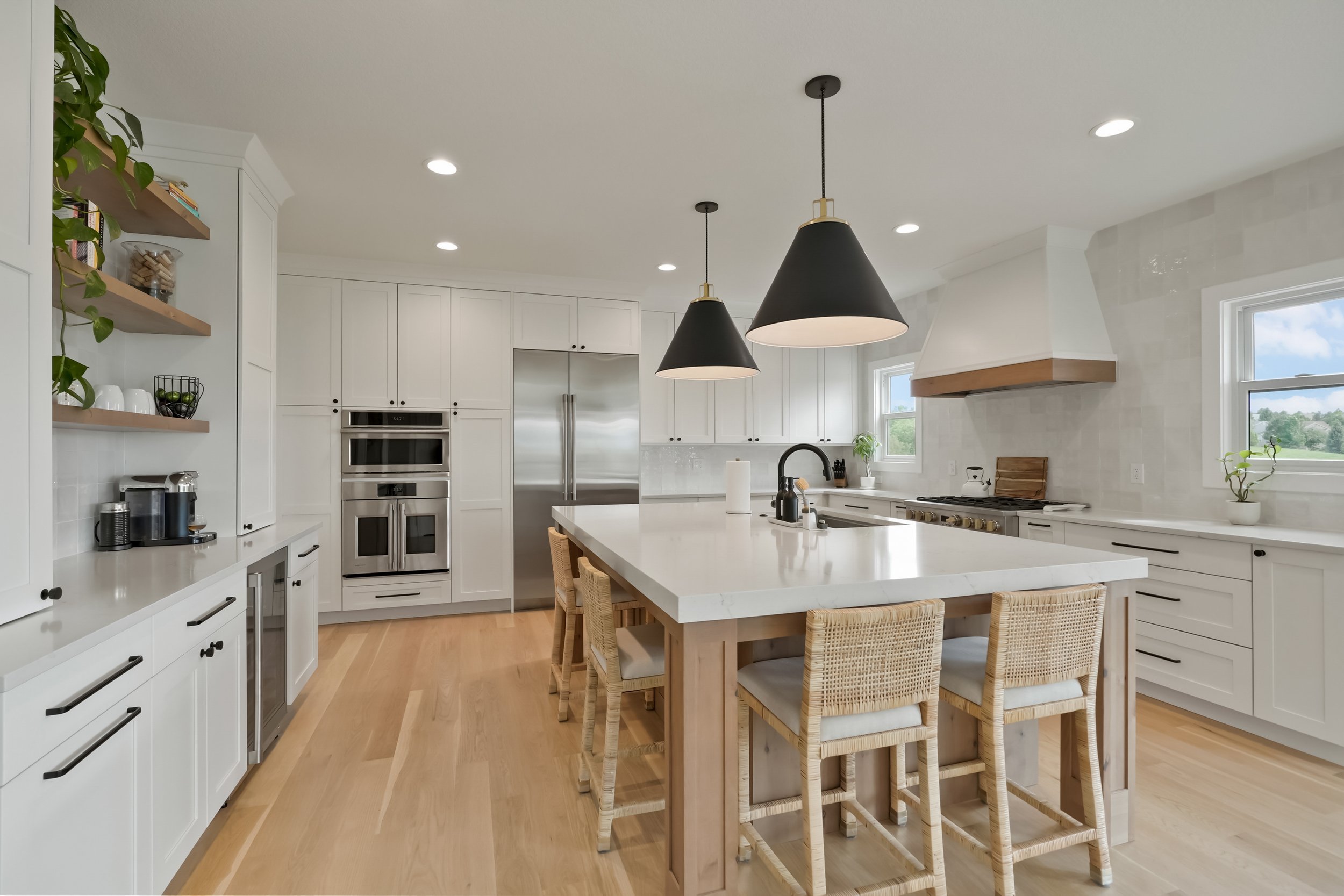Modern white kitchen with island, pendant lights, stainless steel appliances, floating shelves with plants and decor, and large windows showing outdoor greenery.
