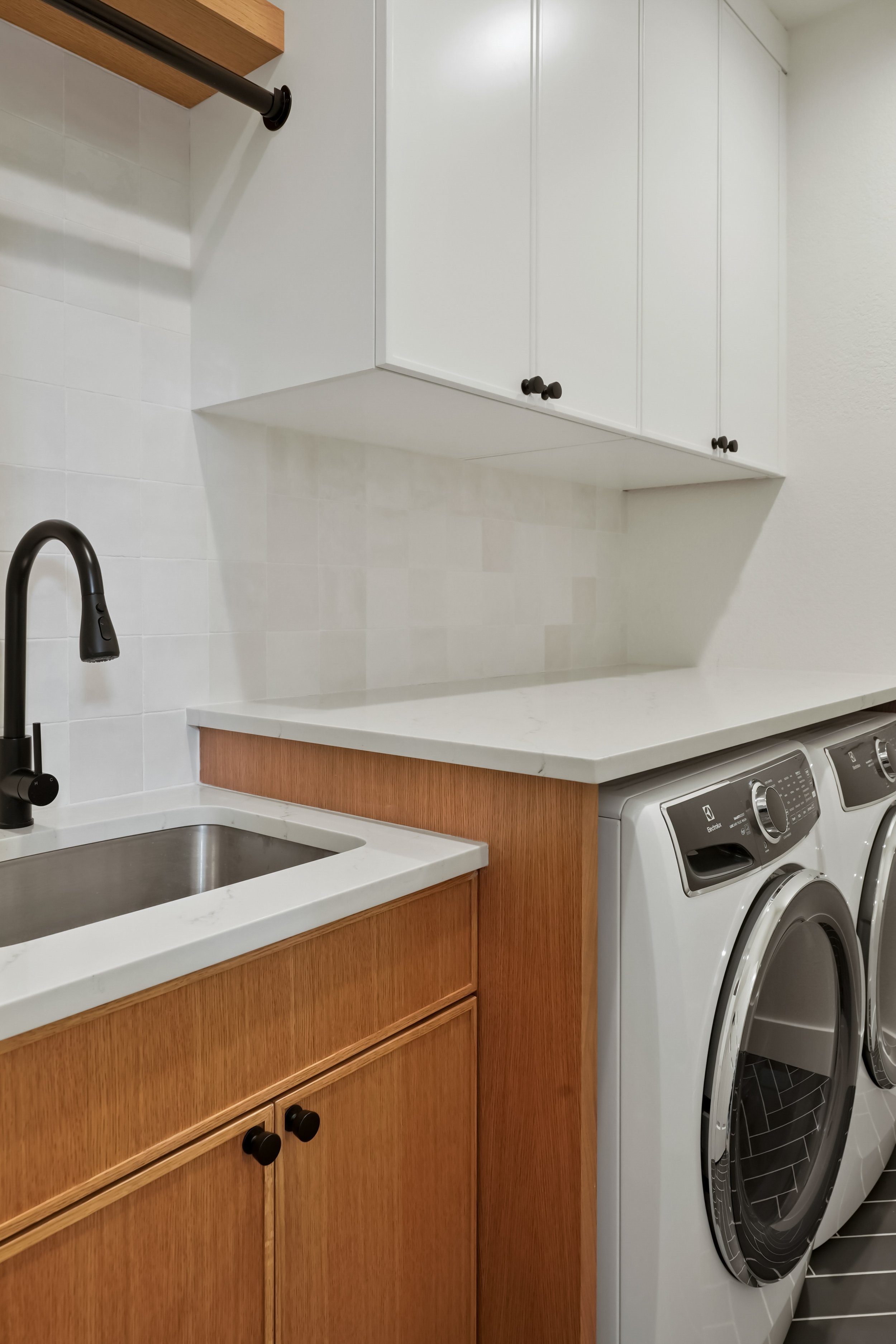Kitchen area with washer and dryer next to a wooden cabinet, white countertop, and a white upper cabinet. The stainless steel sink is visible below the faucet, and the washing machine and dryer are positioned side by side.