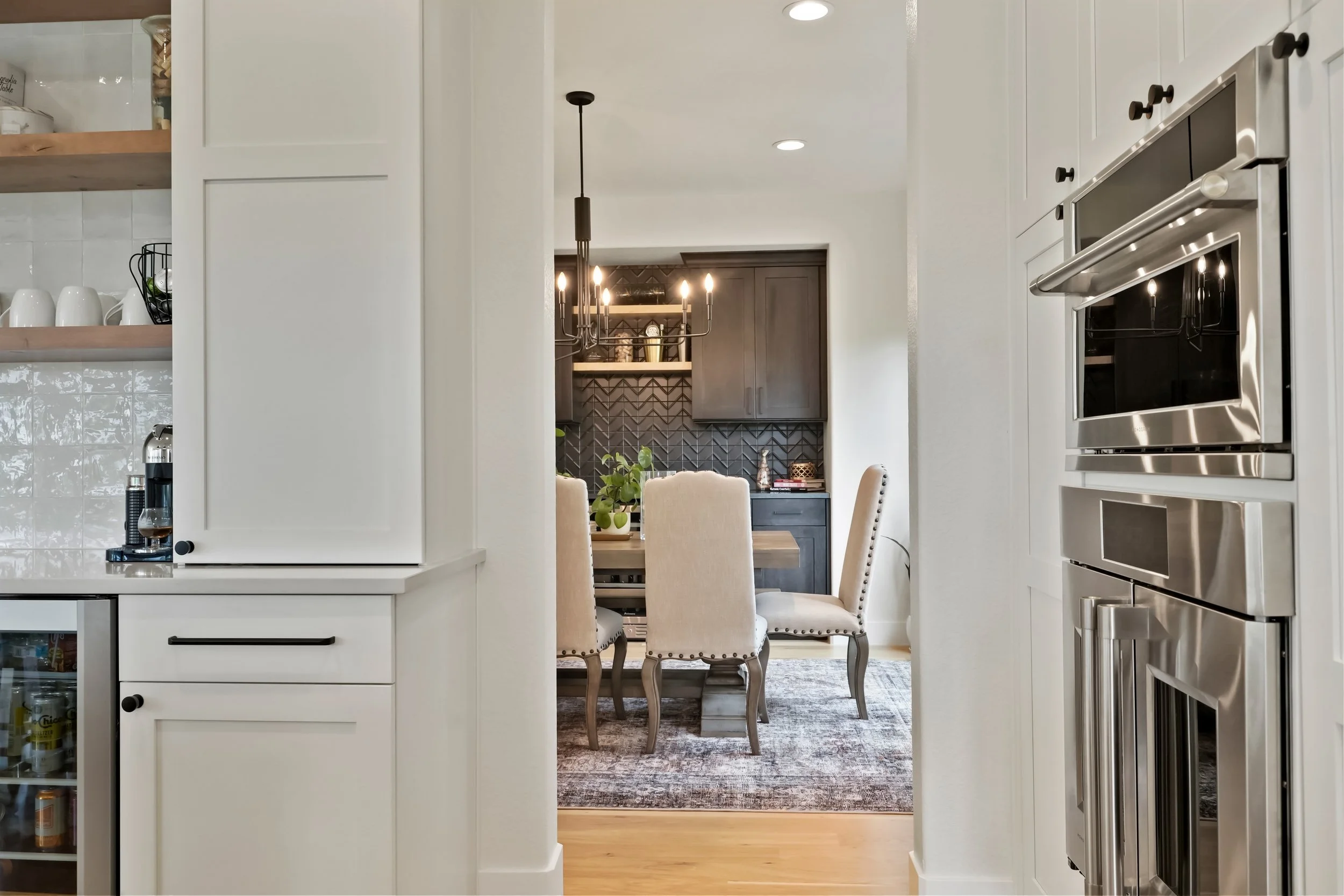 View through a doorway into a dining area with a chandelier, beige upholstered chairs around a wooden table, and a black sideboard against a chevron-patterned wall. Part of a kitchen with white cabinets and built-in stainless steel appliances is visible on either side.