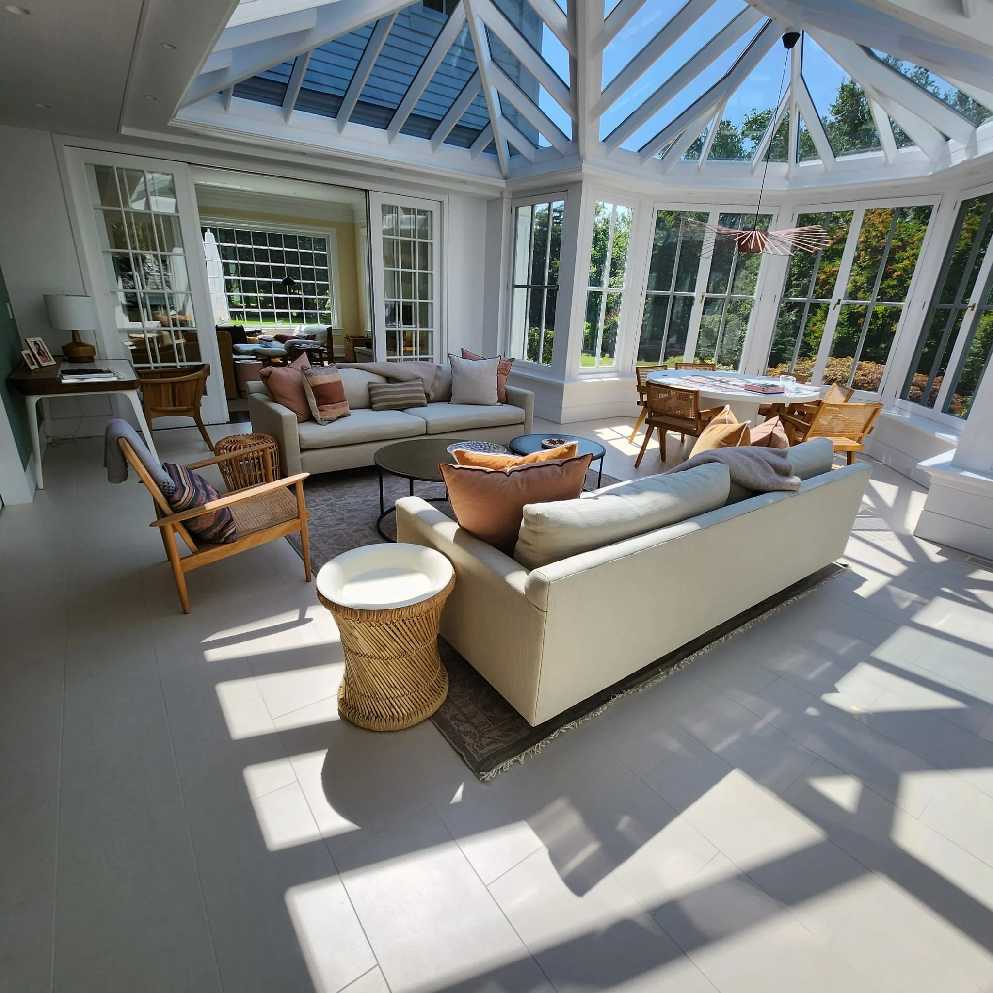 A sunlit glass conservatory with beige sofas, wooden chairs, a rug, and a dining table with chairs, overlooking greenery outside.