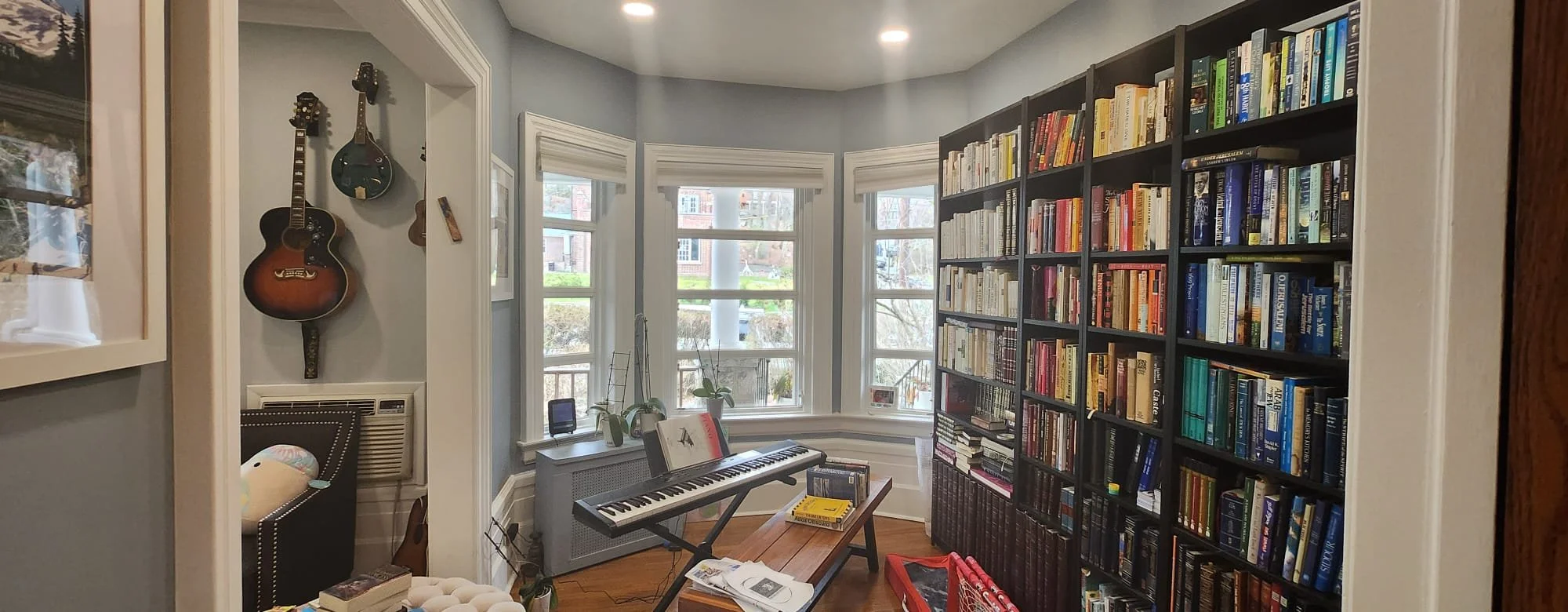View of a cozy reading room with a large bookshelf filled with books, a keyboard on a small table, and three windows letting in natural light.