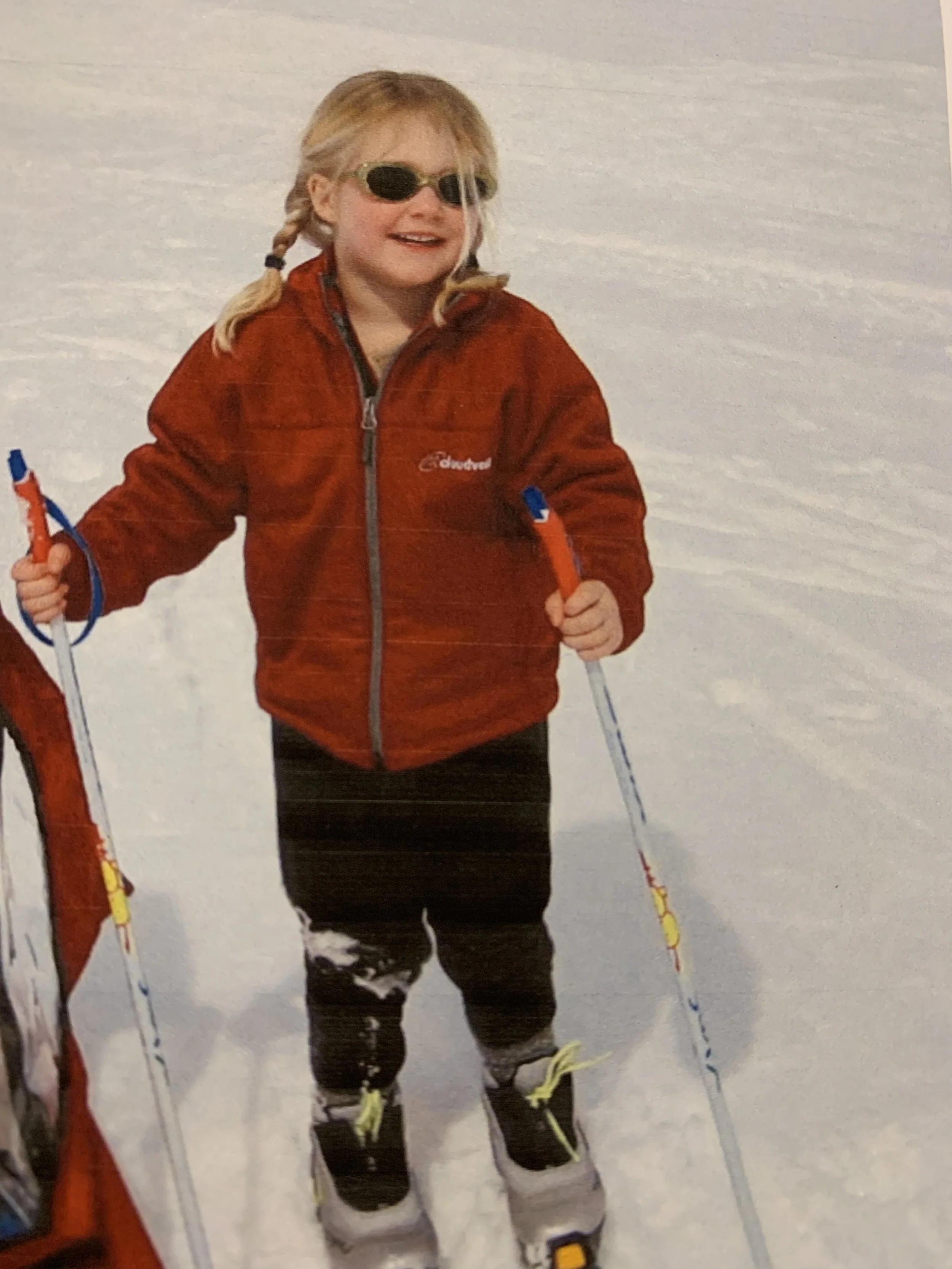 A young girl with blonde hair in pigtails, wearing sunglasses, a red jacket, black pants, and ski boots, holding ski poles on a snowy landscape.