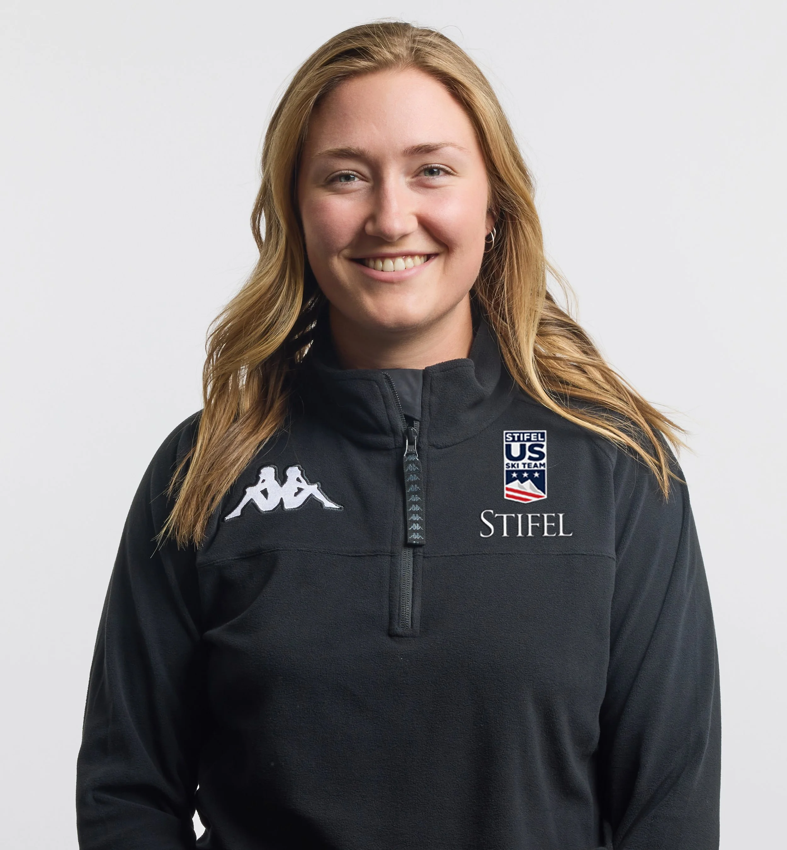 Young woman with long blonde hair smiling, wearing a black zip-up sports jacket with sports logos on the chest.