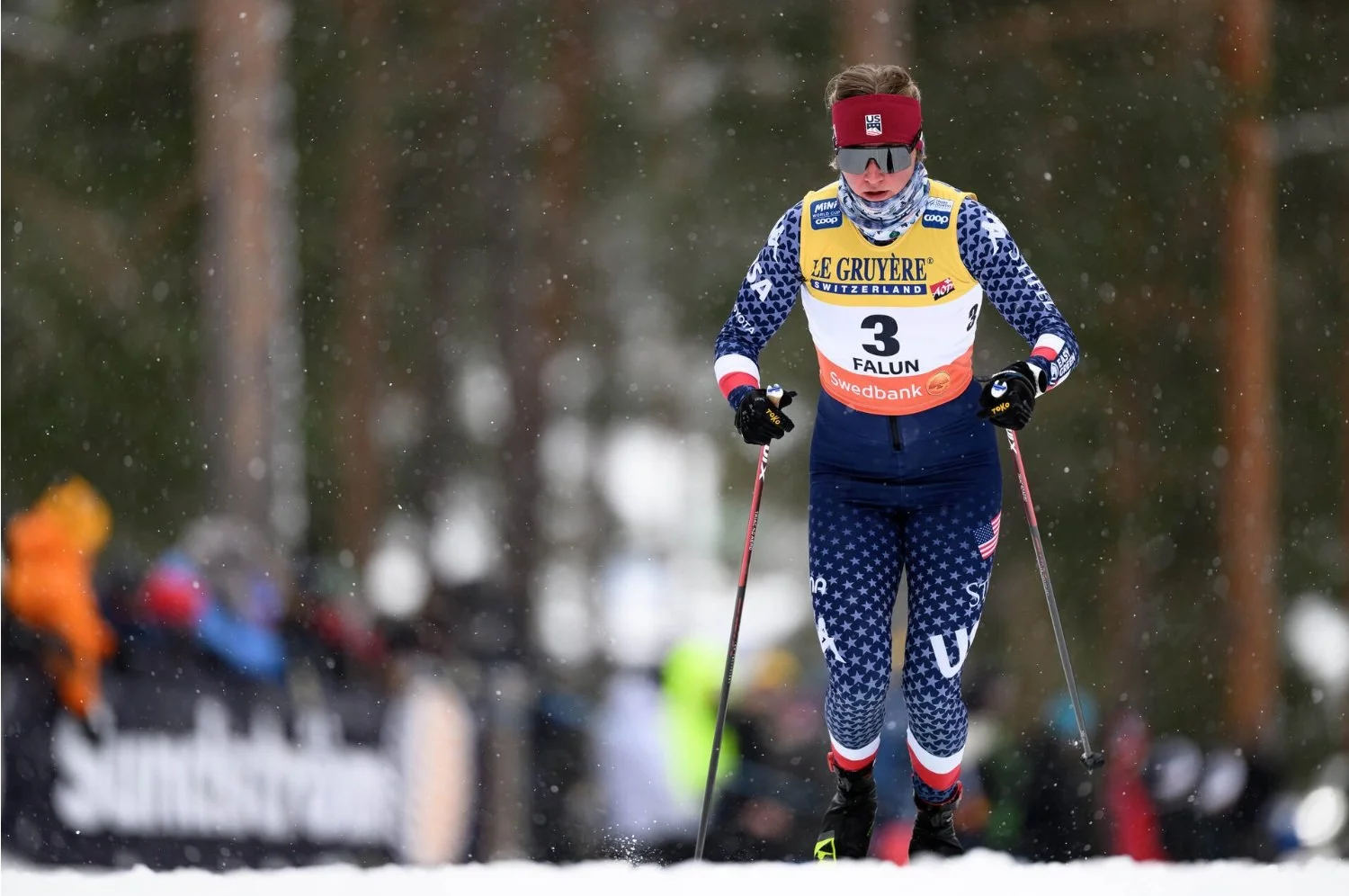 A cross-country skier from the United States racing on snow in winter, wearing a red headband, dark sunglasses, a patriotic blue star-patterned suit, and a yellow bib with the number 3, with a forest background.