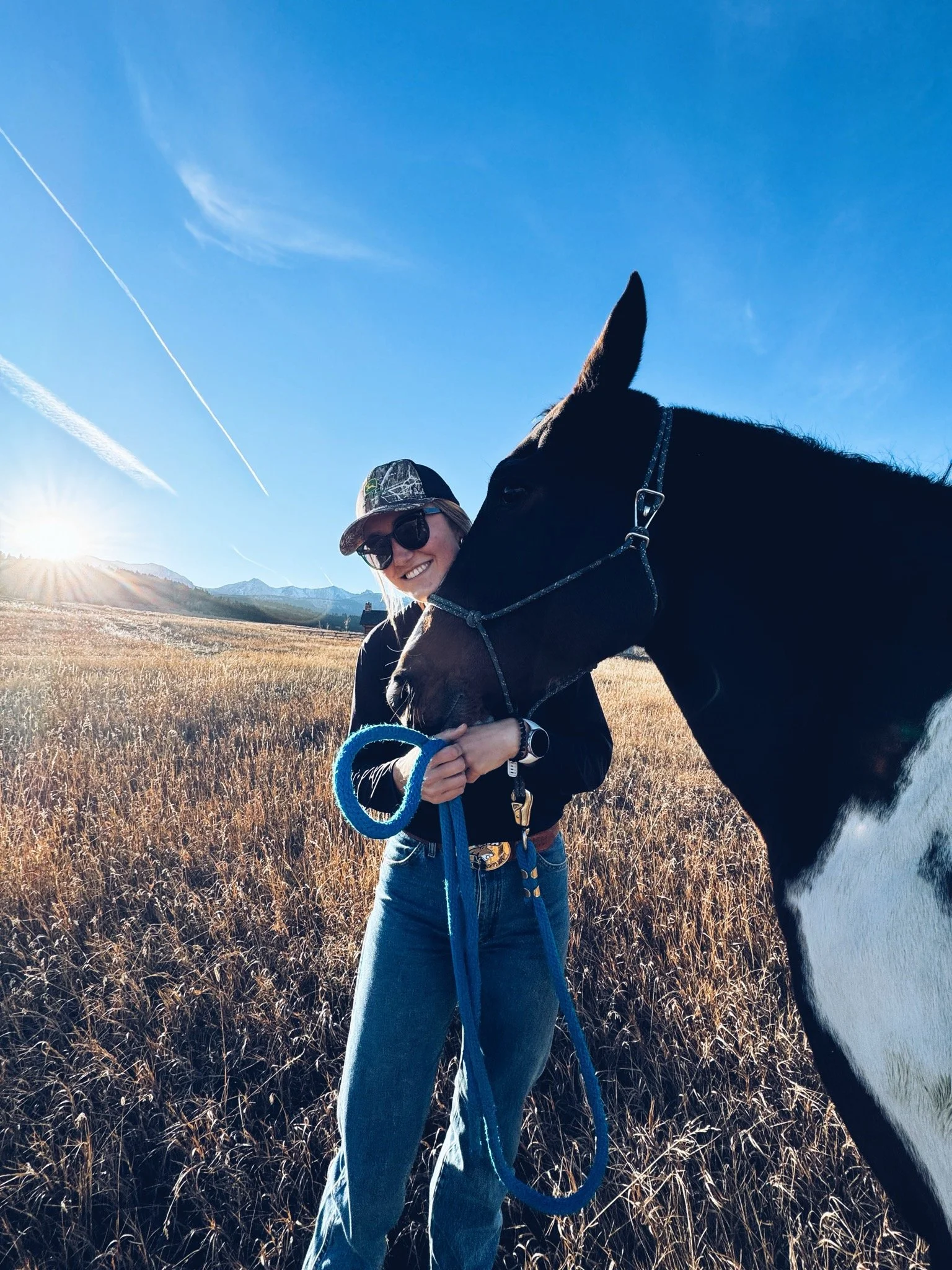 A woman smiling and holding a blue leash next to a black and white horse in a grassy field with mountains and a clear blue sky in the background during sunset.