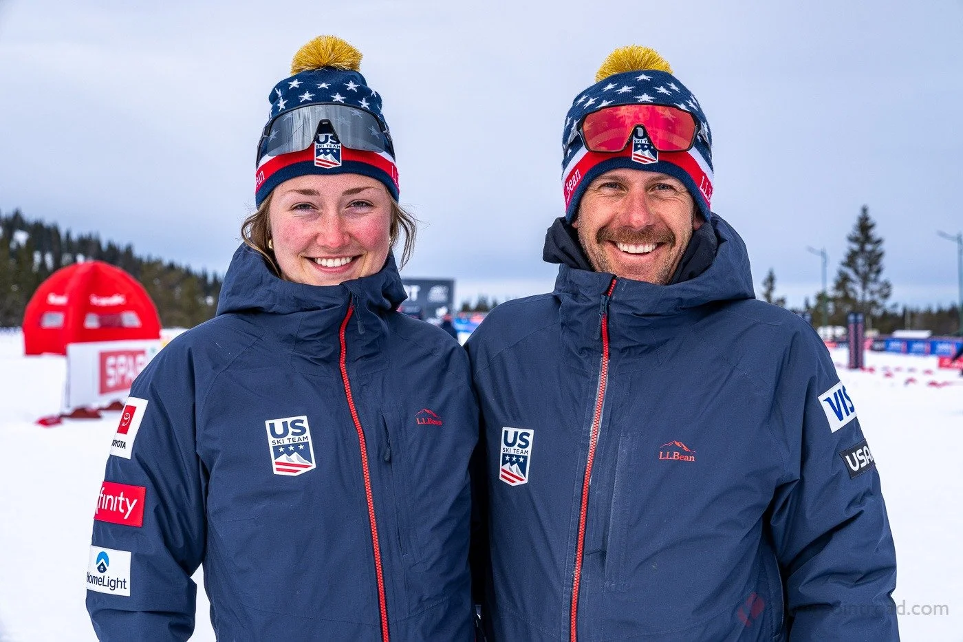 Two skiers in blue USA Ski Team jackets smiling outdoors in snowy setting, both wearing USA-themed beanies with pom-poms, ski goggles, and jackets with various sponsor patches.