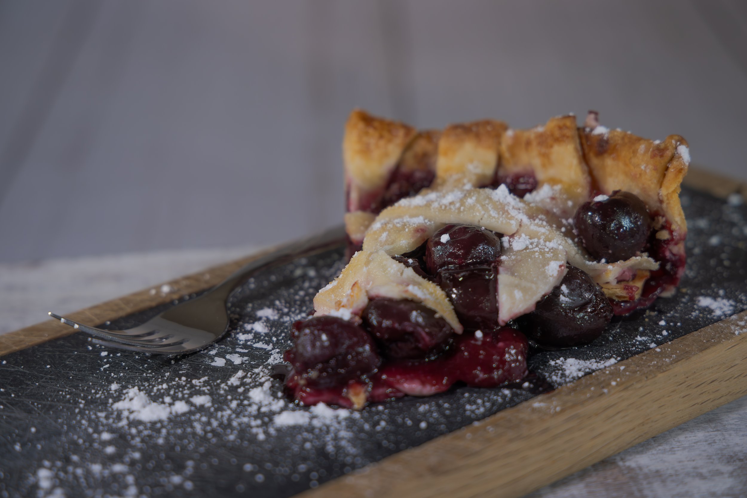 Slice of cherry pie with powdered sugar on a slate plate, fork beside it.