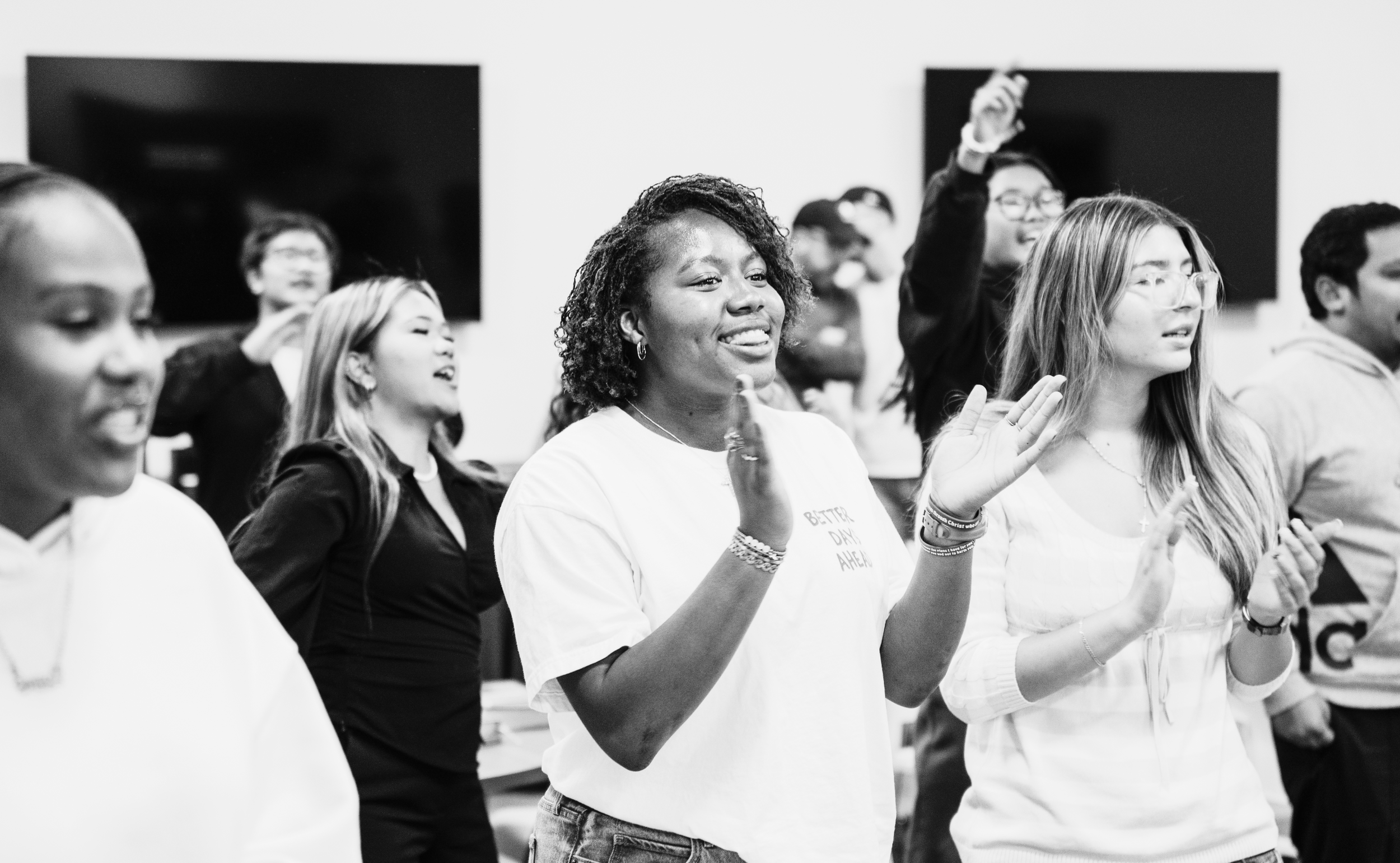 Black and white photo of a diverse group of people at an indoor event, smiling and engaging, with two large black screens or monitors in the background.