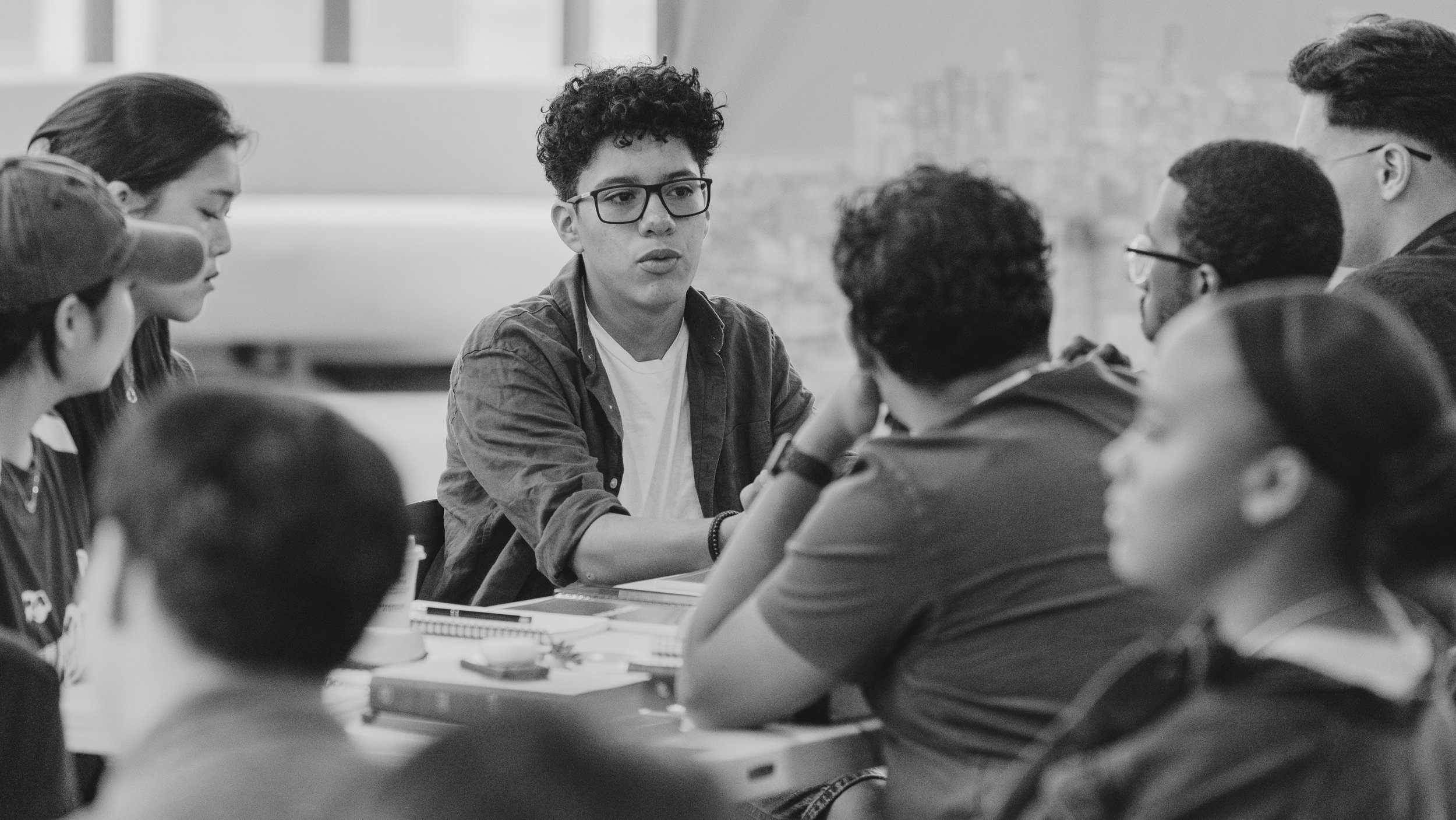 Group of young people sitting around a table engaged in conversation in a classroom or meeting room.