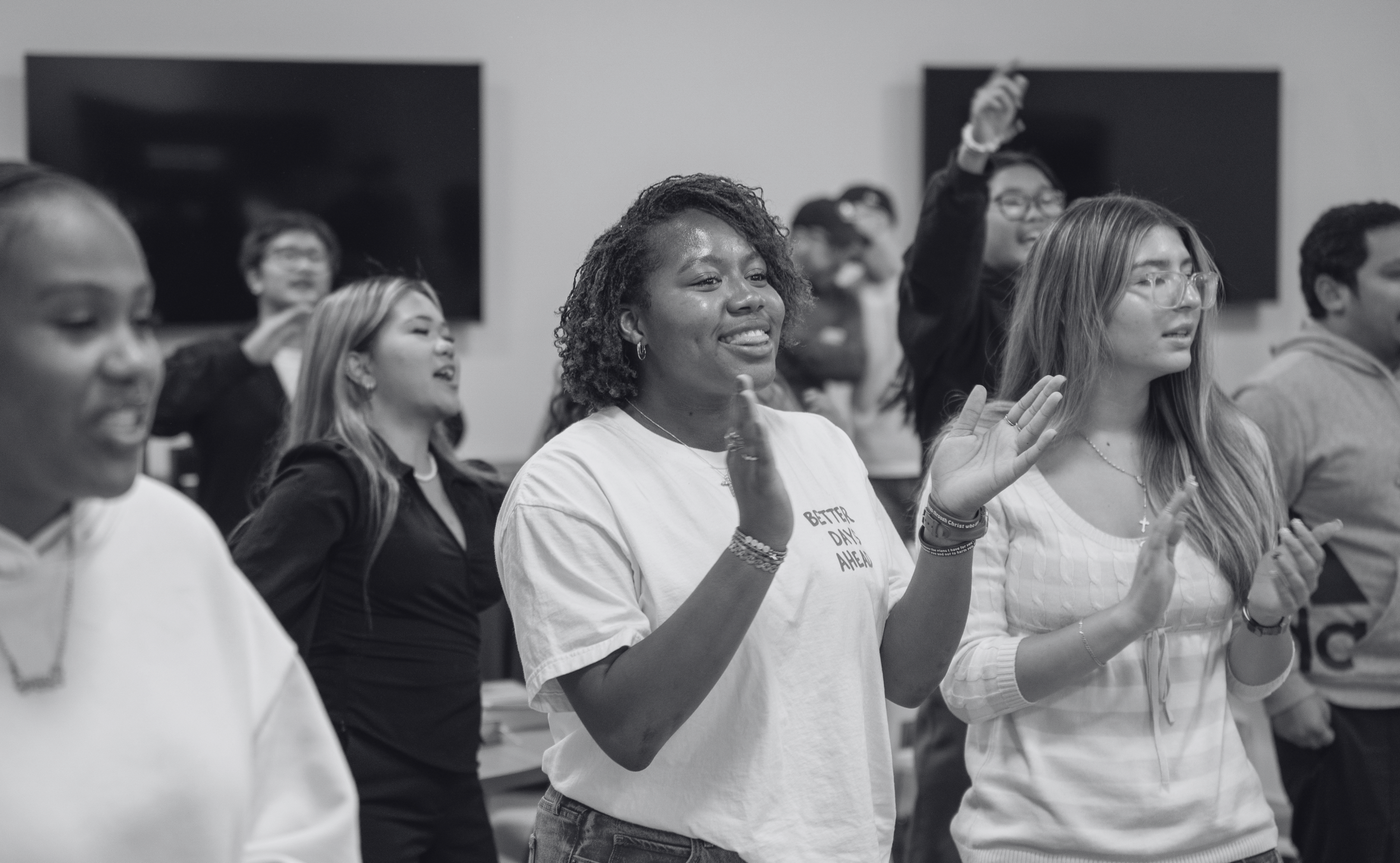 A diverse group of people singing or participating in a group activity, some with their eyes closed or smiling, with a woman in the center clapping her hands and wearing a shirt that says 'Better Day Ahead.'
