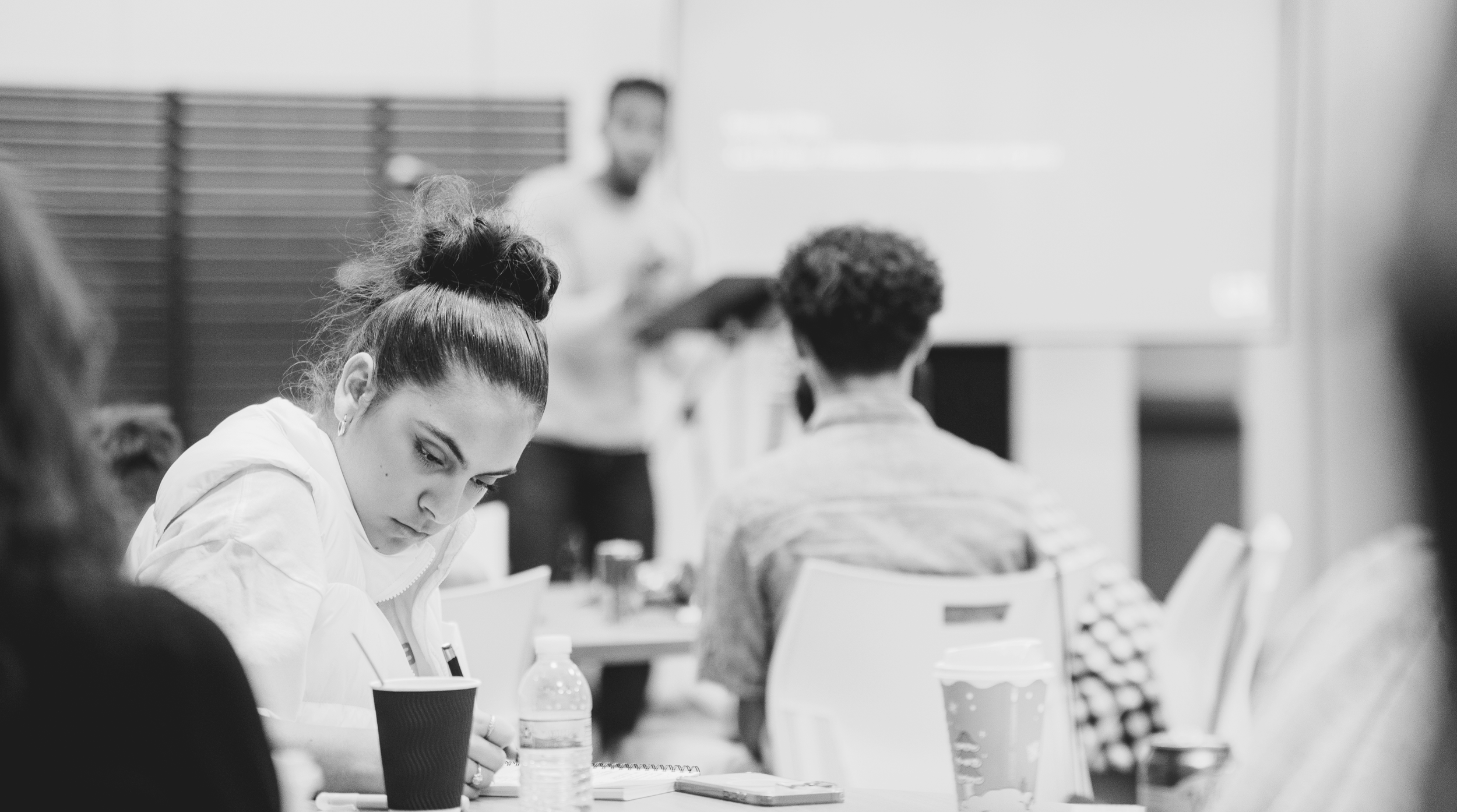 People attending a meeting or conference in a classroom setting, with a woman in the foreground taking notes.