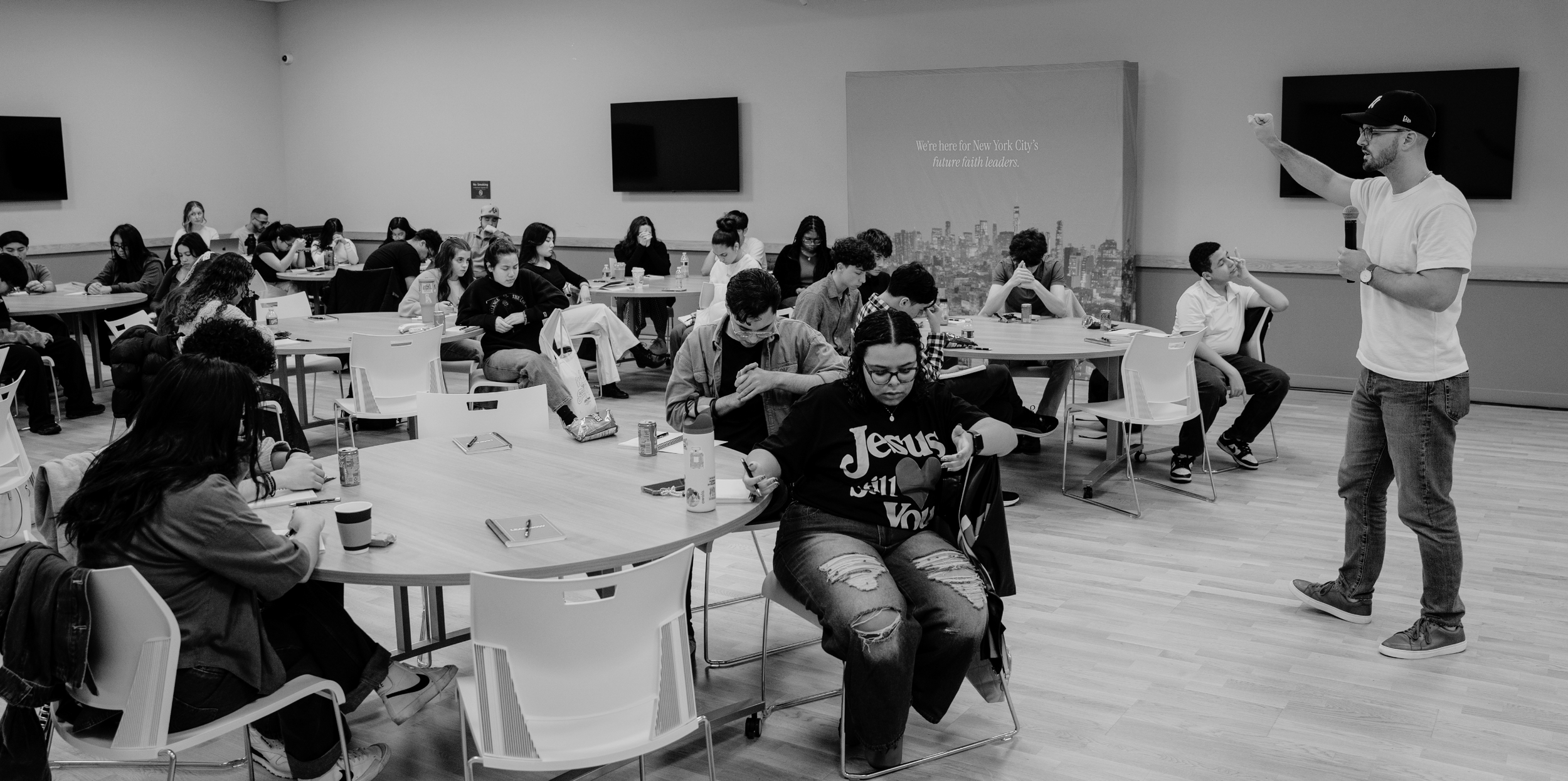 A group of young people seated at round tables in a classroom or seminar room, listening to a man standing at the front with a microphone, giving a presentation or lecture. The room has a large cityscape backdrop and two wall-mounted screens.