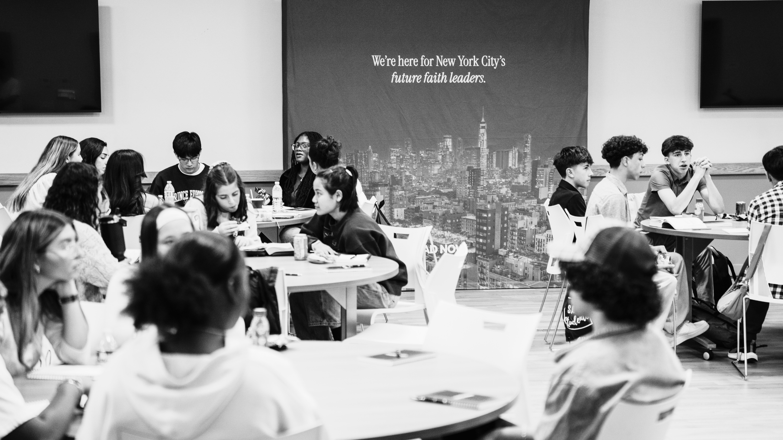 A group of young people sitting around tables in a modern room with a large cityscape poster and screens on the walls.