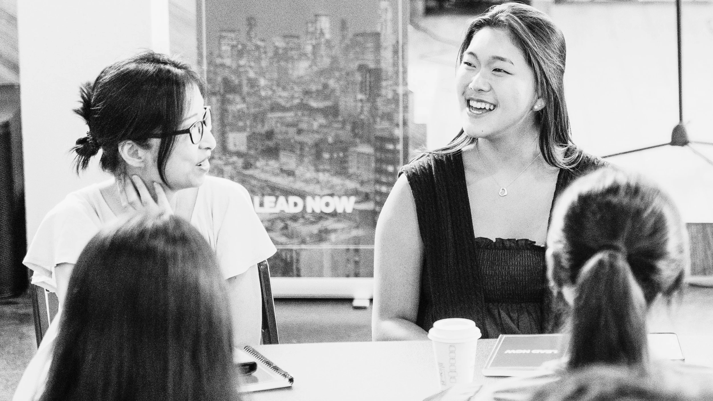 Two women, one wearing glasses, engaged in conversation in an indoor setting with a cityscape view in the background, and several people partially visible in the foreground.