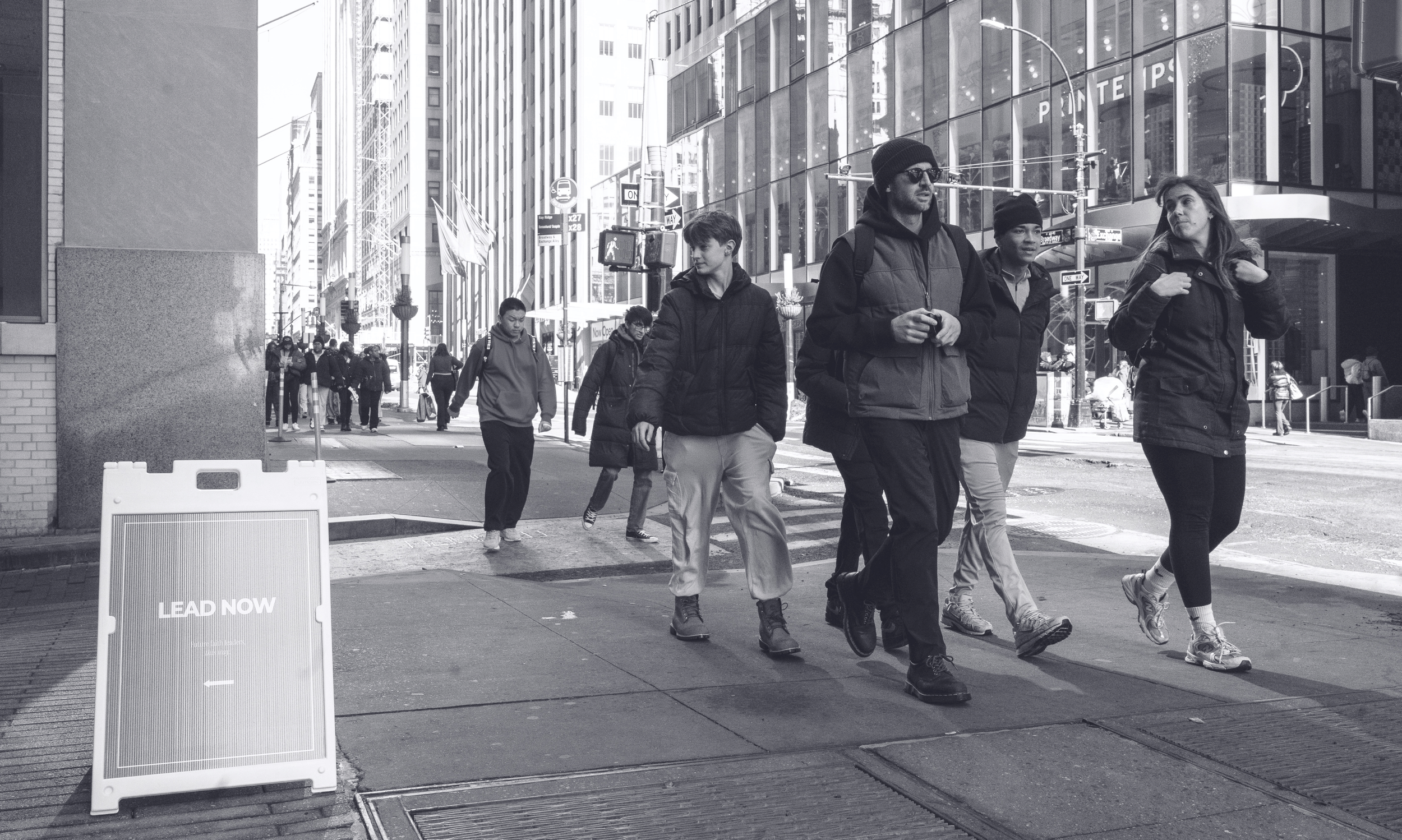 A group of diverse pedestrians crossing a city street, with tall buildings and glass windows in the background. There is a sign on the sidewalk that says 'LEAD NOW'.