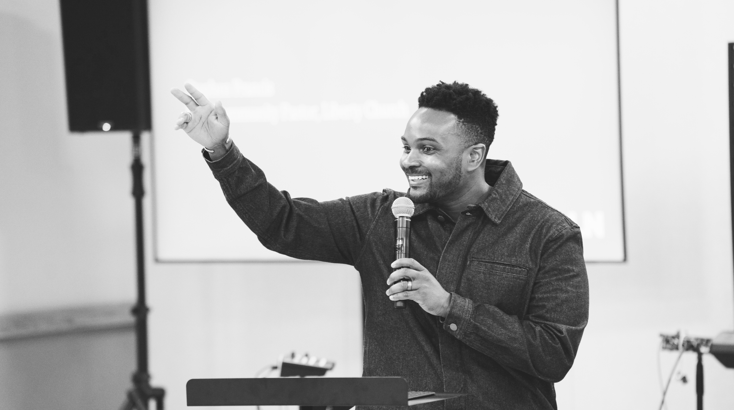 A man with a beard and short curly hair holding a microphone and gesturing with his left hand while speaking at an indoor event.