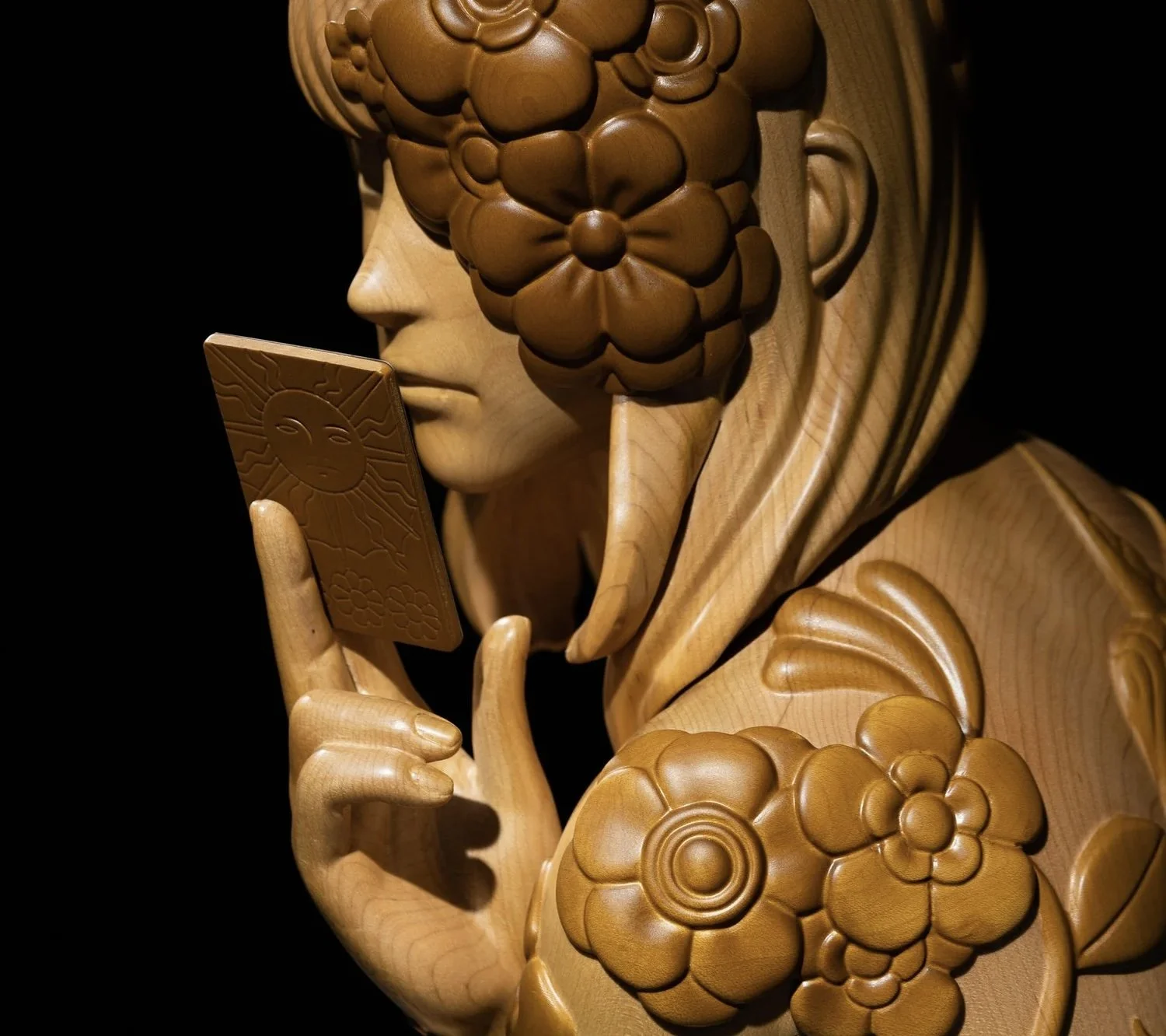 A wooden sculpture of a person with detailed floral and facial features, holding a phone decorated with a sun and face design, all lit against a dark background.