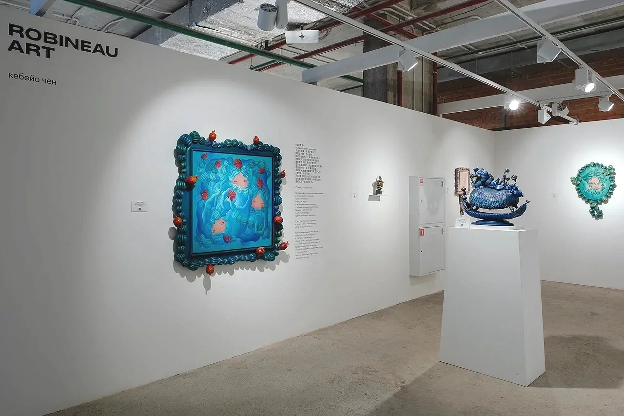 An art gallery display featuring colorful sculptures, including a blue framed piece with cartoonish figures surrounded by a border of rounded objects, a blue ornate vessel sculpture on a pedestal, and a green decorative wall hanging on a white wall.