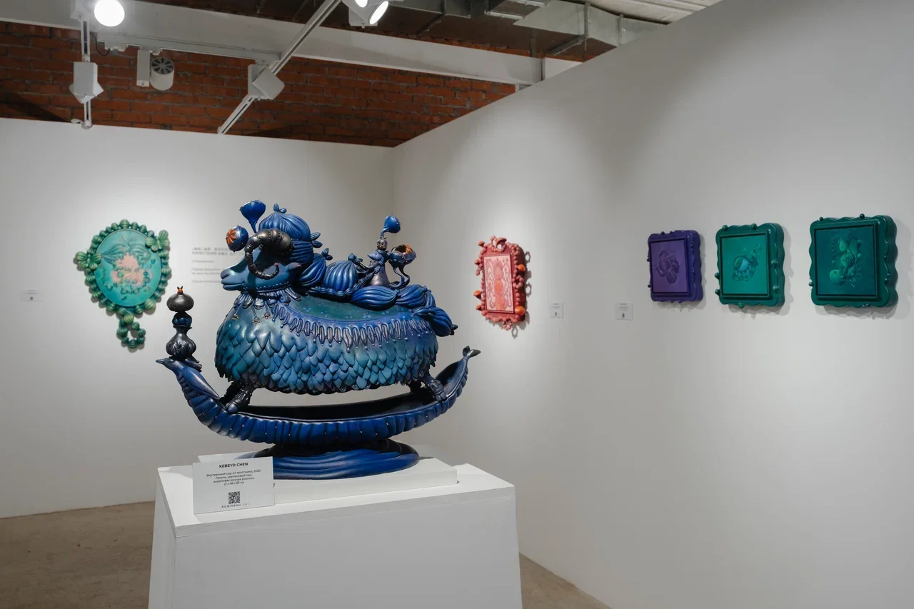 Colorful art exhibition featuring a large blue sculpture resembling a mythical creature and framed artworks on a white gallery wall.