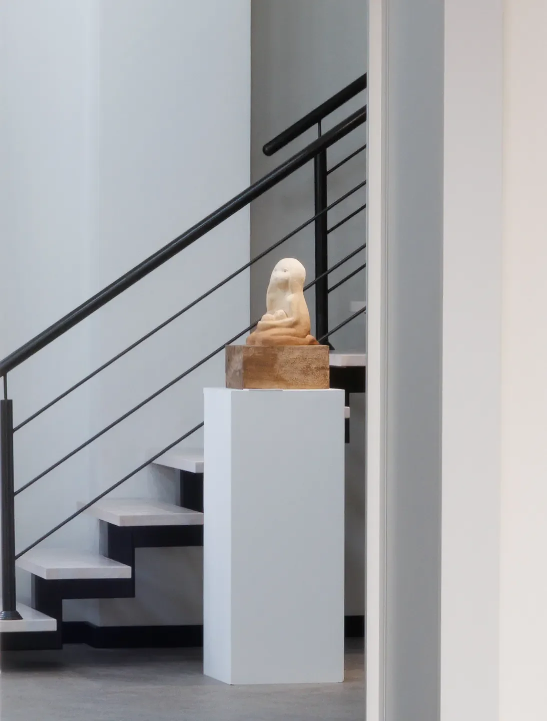 A sculpture of an abstract, rounded figure sits on a wooden pedestal, positioned on a white display stand near a staircase in an art gallery.