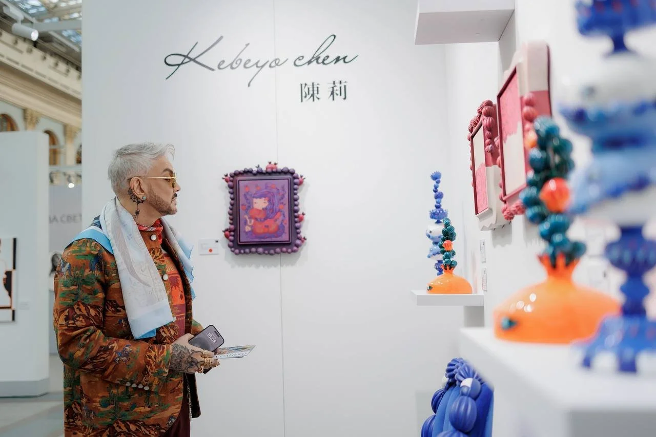 An elderly man with gray hair, sunglasses, and tattoos is looking at colorful glass sculptures in an art gallery. The gallery has white walls with artwork and the name 'Kebeya chen' written on the wall.