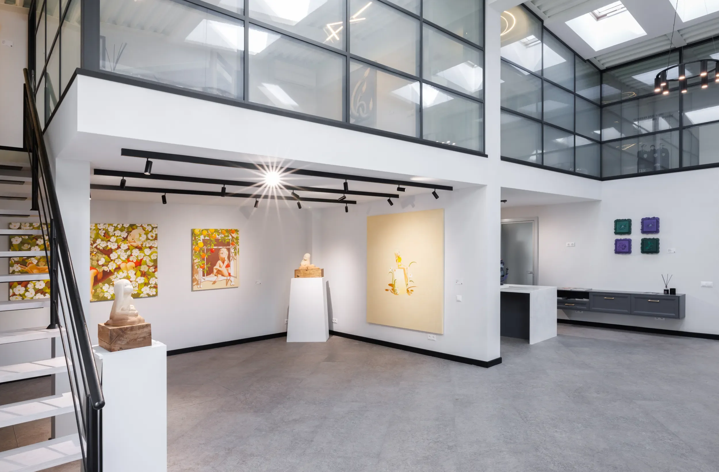 Interior view of an art gallery with paintings and sculptures on display, modern decor, large glass windows, and a staircase to the left.