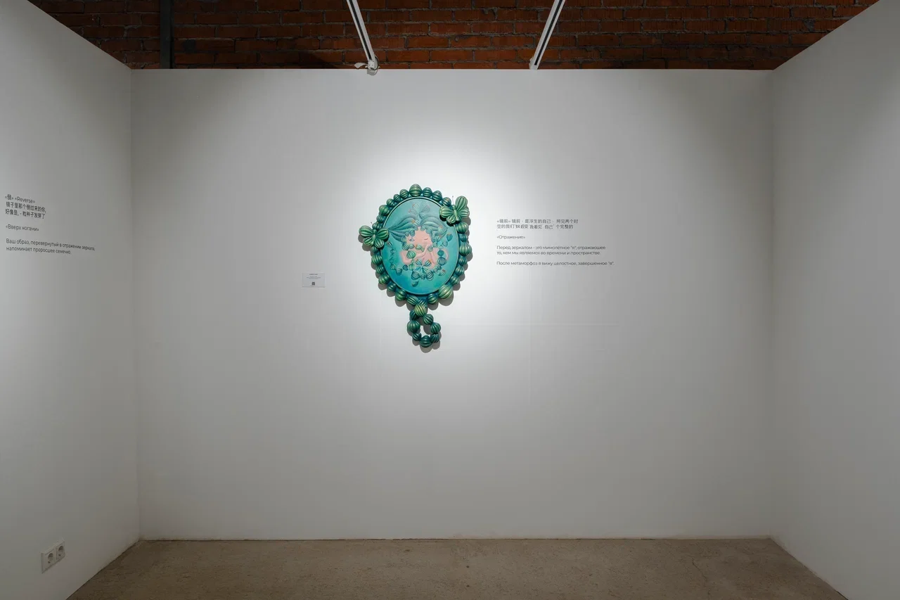 Art installation of a decorated mirror with a blue-green fish and floral design on white gallery wall.