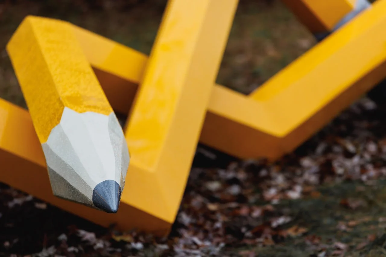 A close-up of a yellow wooden pencil with a white eraser tip resting on a yellow wooden bench outdoors, with fallen leaves on the ground.