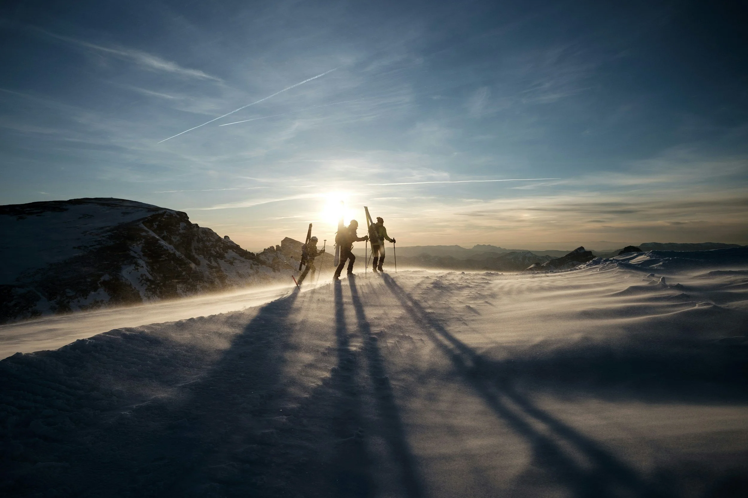 Three hikers walking on a snowy mountain trail at sunset, with long shadows cast on the snow and a mountain range in the background.