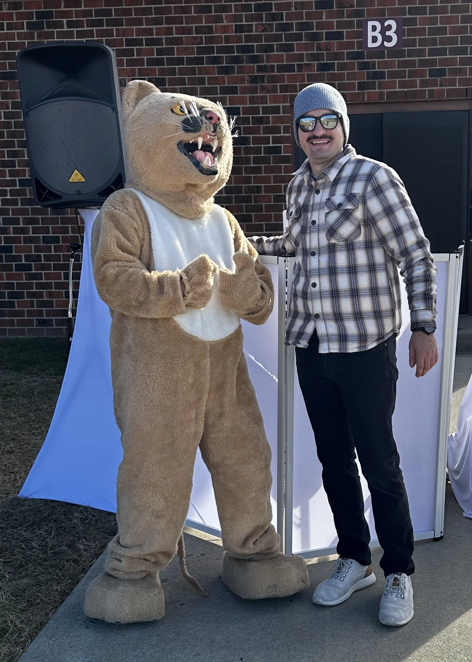 Person in a plaid shirt and beanie standing next to a person in a lion mascot costume outdoors in front of a brick building.