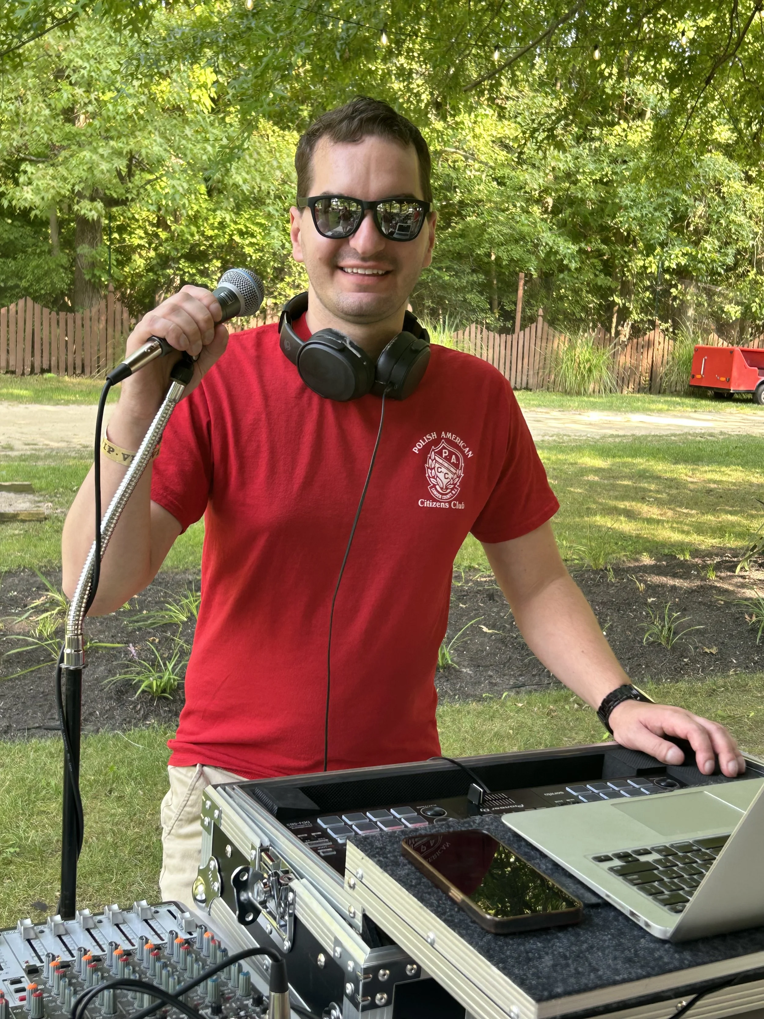 A man holding a microphone and wearing headphones, standing outdoors with DJ equipment and a laptop, smiling at the camera during daytime.