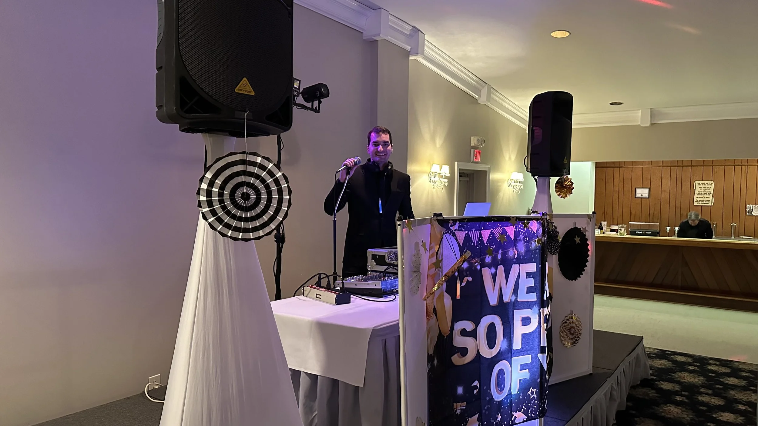 A man dressed in a black jacket stands behind a DJ booth at a party, smiling and holding a microphone. The booth is decorated with festive banners and black, white, and gold decorations. Large speakers are positioned on either side of the booth, and 