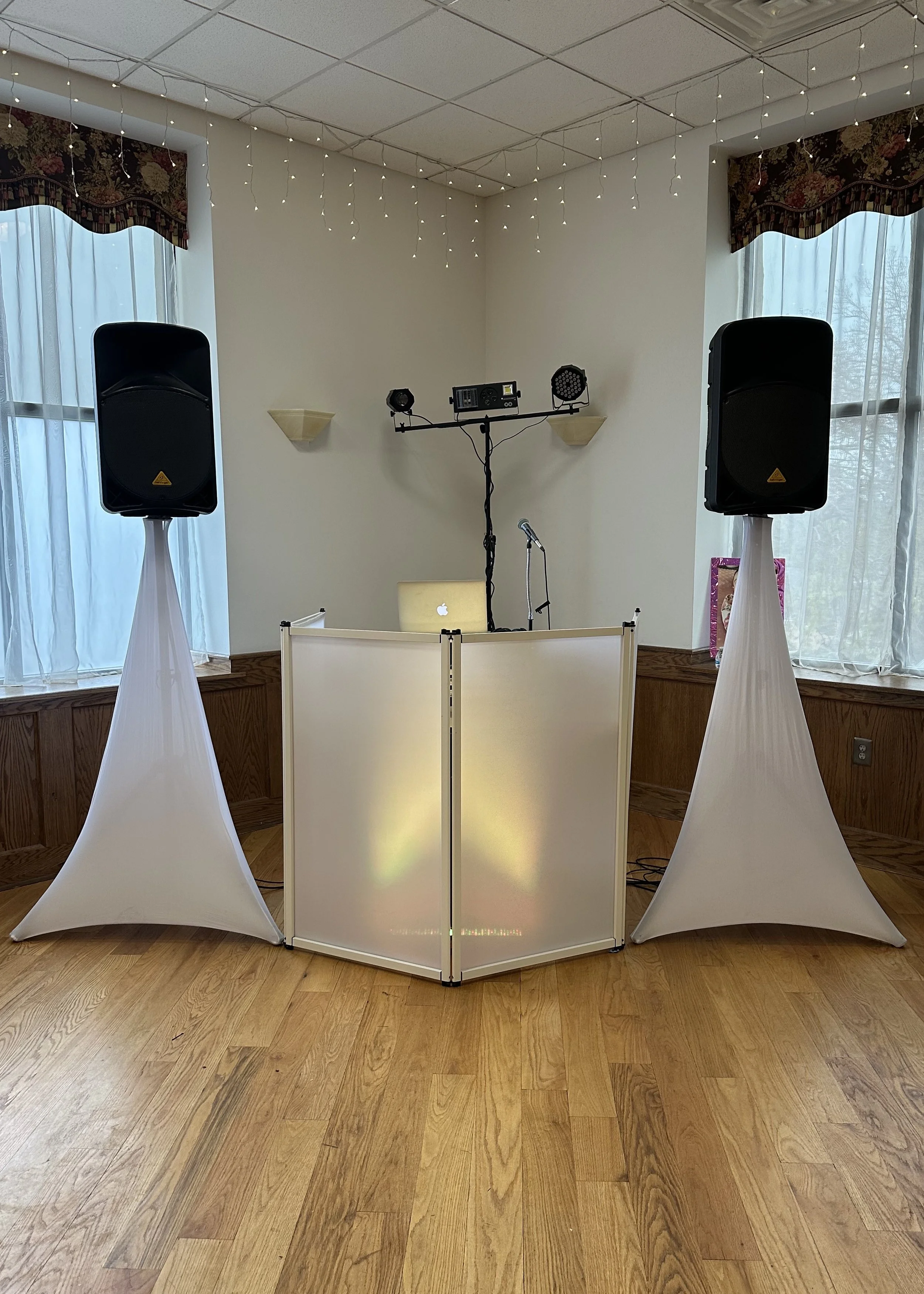 DJ setup with two large black speakers on stands, a white DJ booth with a frosted panel, a laptop, and lighting equipment on a stand in a room with hardwood floors and windows with sheer curtains.
