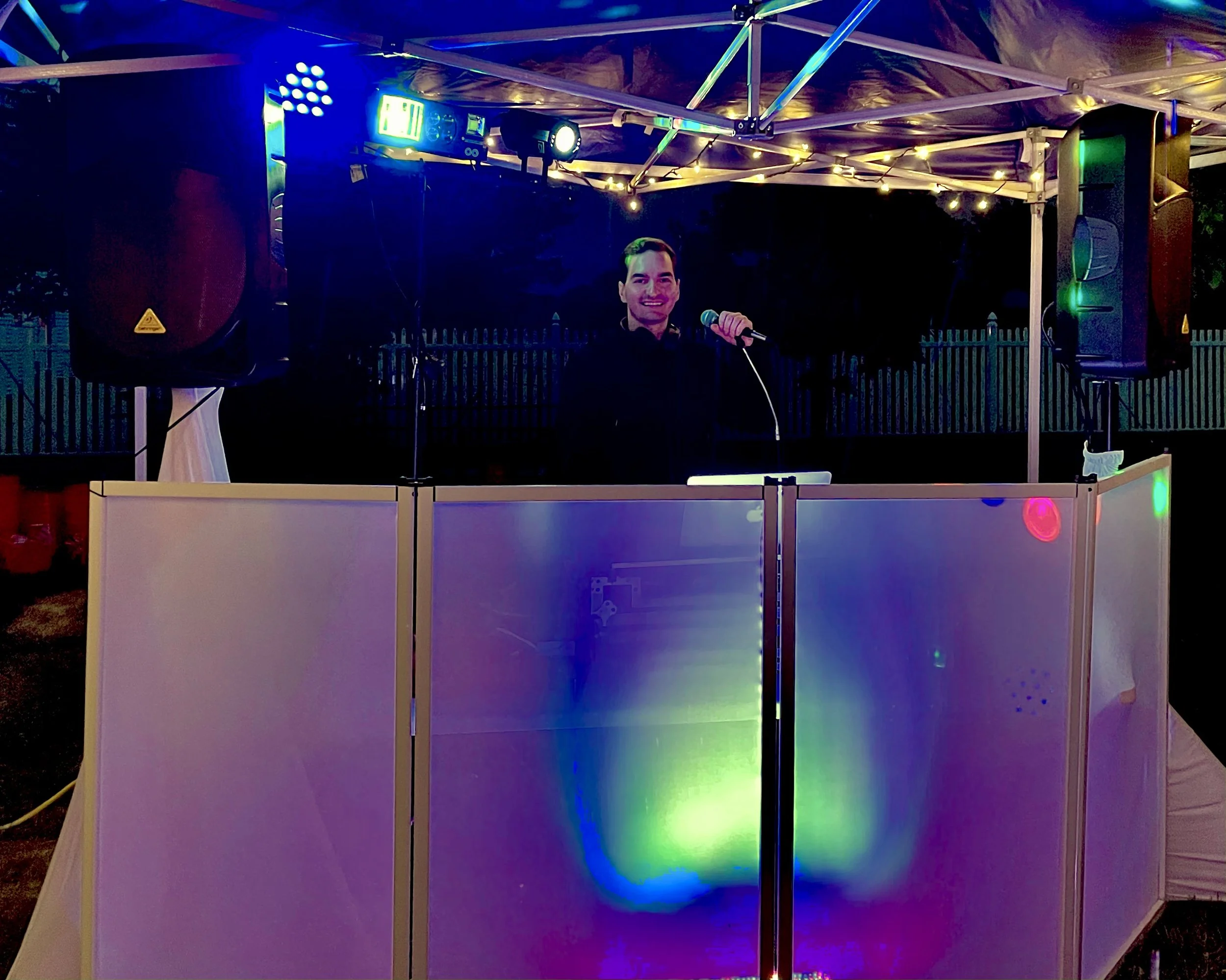 A man is smiling and speaking into a microphone at a nighttime outdoor event, standing behind a white DJ booth with colorful LED lights, under a canopy with string lights. Large speakers are positioned on either side of him, with a dark sky and a fen