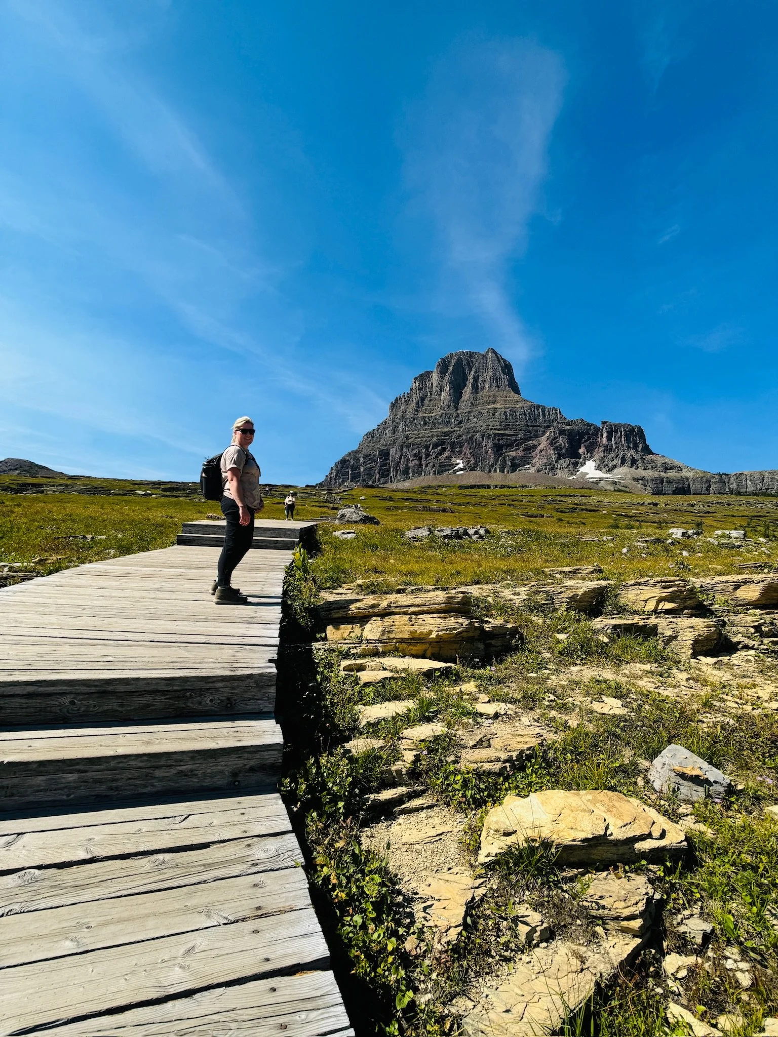 Hiker walking on a wooden trail in a mountainous landscape with a large rocky mountain in the background, under a clear blue sky.