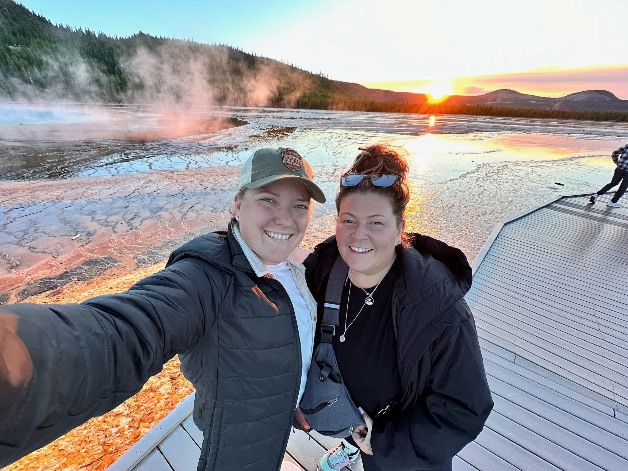 Two women smiling and taking a selfie on a wooden boardwalk at sunset, with hot springs and steam rising from the water in the background.