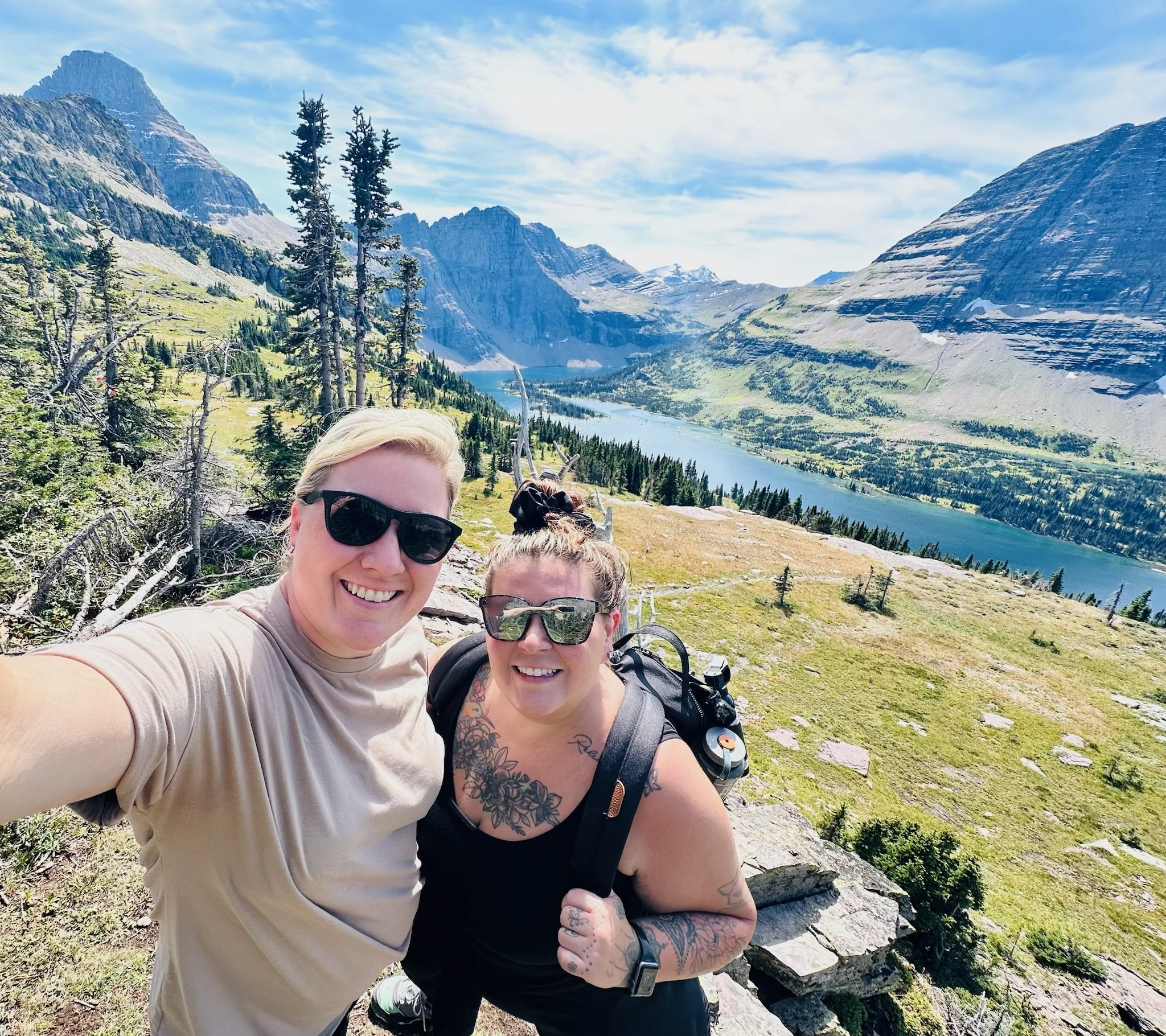 Two women hiking on a mountain trail with a scenic view of a lake and mountains in the background.