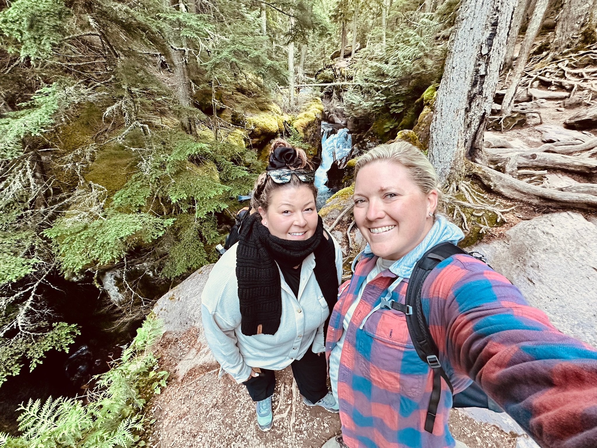 Two women backpacking in a forest, taking a selfie on a trail next to a small waterfall surrounded by tall trees and green moss.